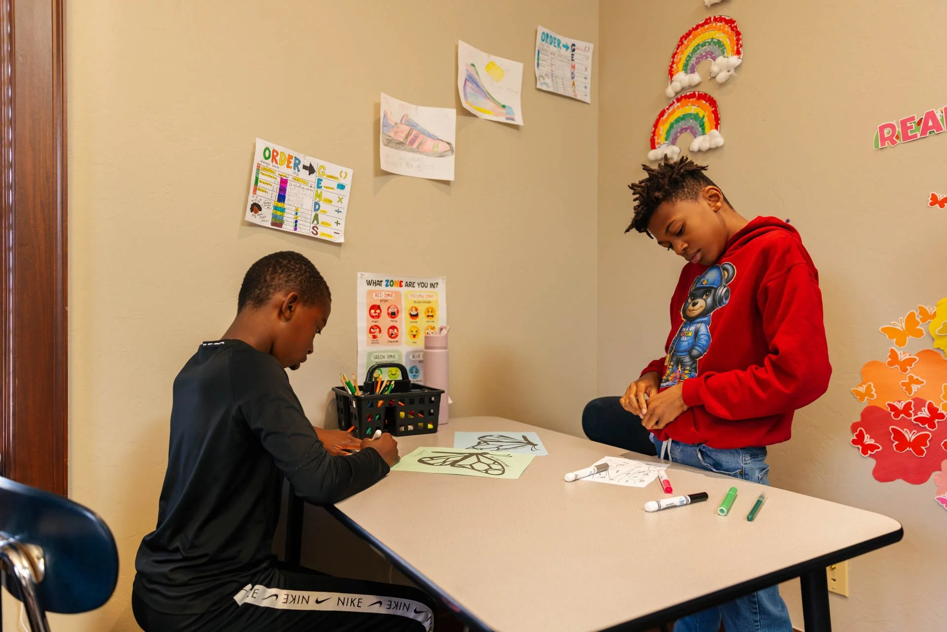 Two boys in a classroom, one sitting at a table drawing and the other standing, wearing a red hoodie with a cartoon character. The wall behind them has colorful posters, drawings, and rainbow decorations.