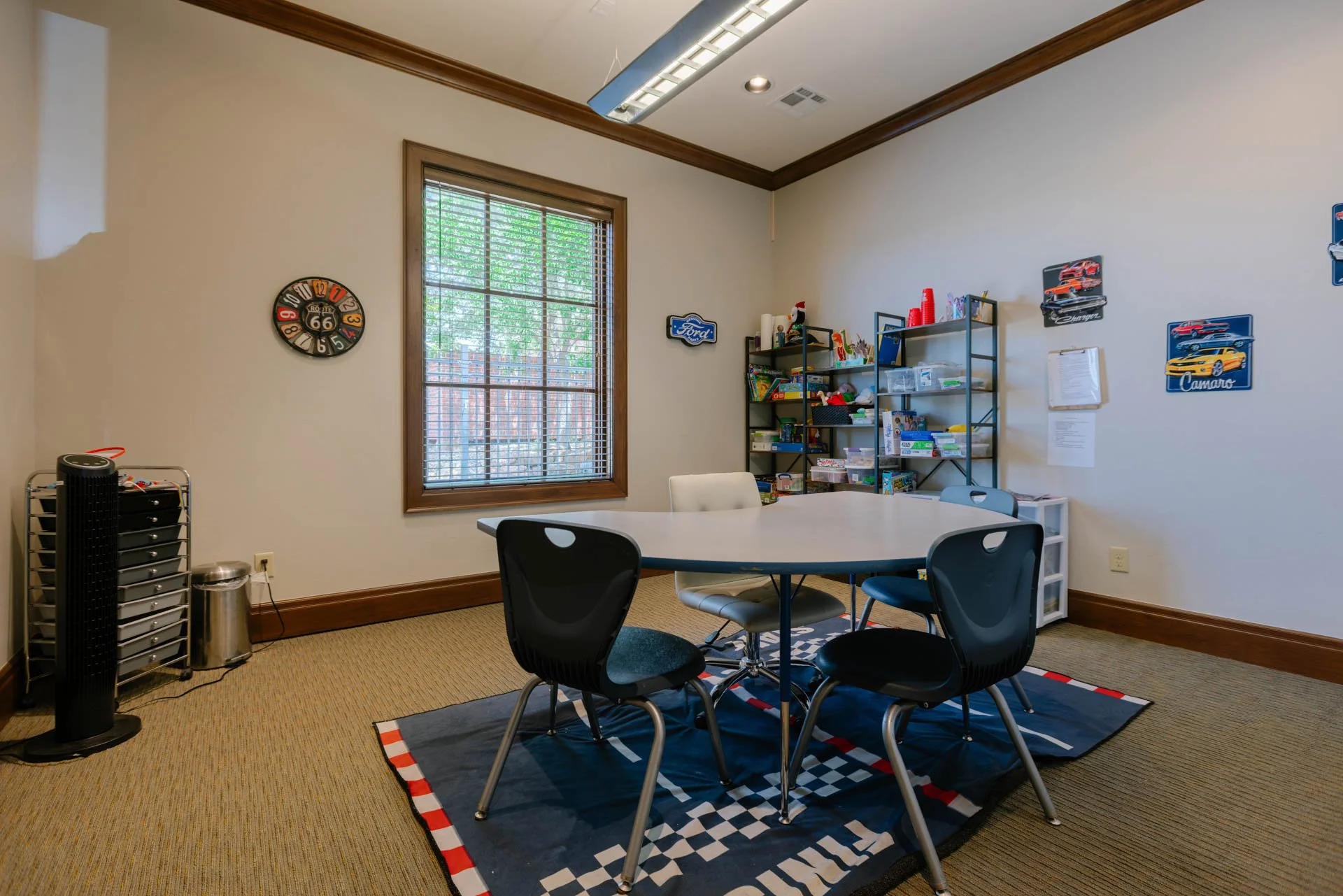 A meeting or break room with a round table and four chairs, decorated with a racing flag-themed rug. There are shelves with toys and books, a window with blinds, and automobile-themed decorations on the wall. A wall clock shaped like a race car wheel and a black tower fan are also visible.