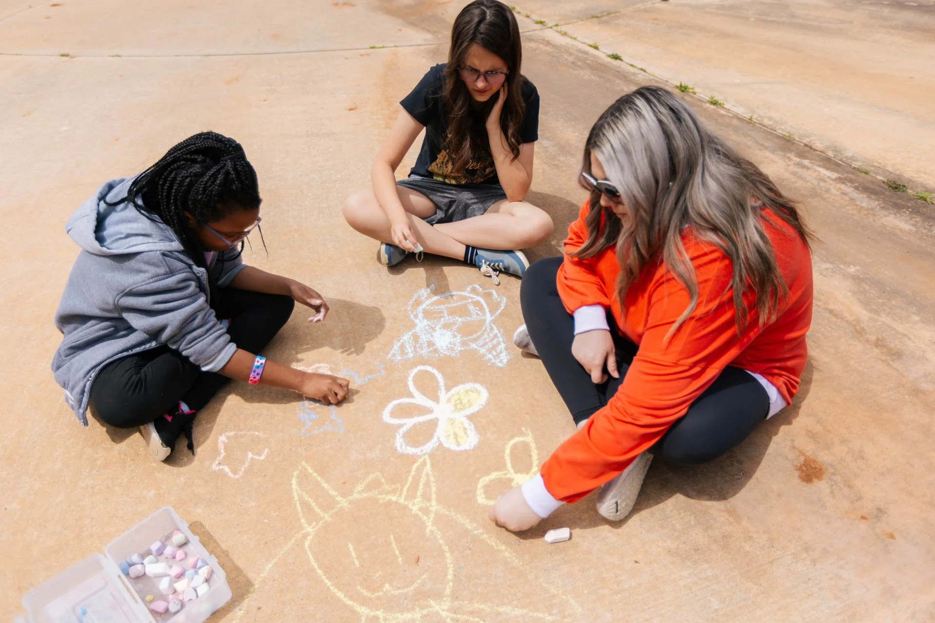 Three girls drawing and coloring on sidewalk with chalk, surrounded by pastel chalks in a container, on a sunny day.