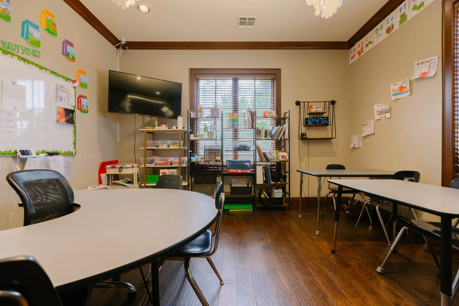 A classroom with tables and chairs, decorated with educational posters and books on shelves.