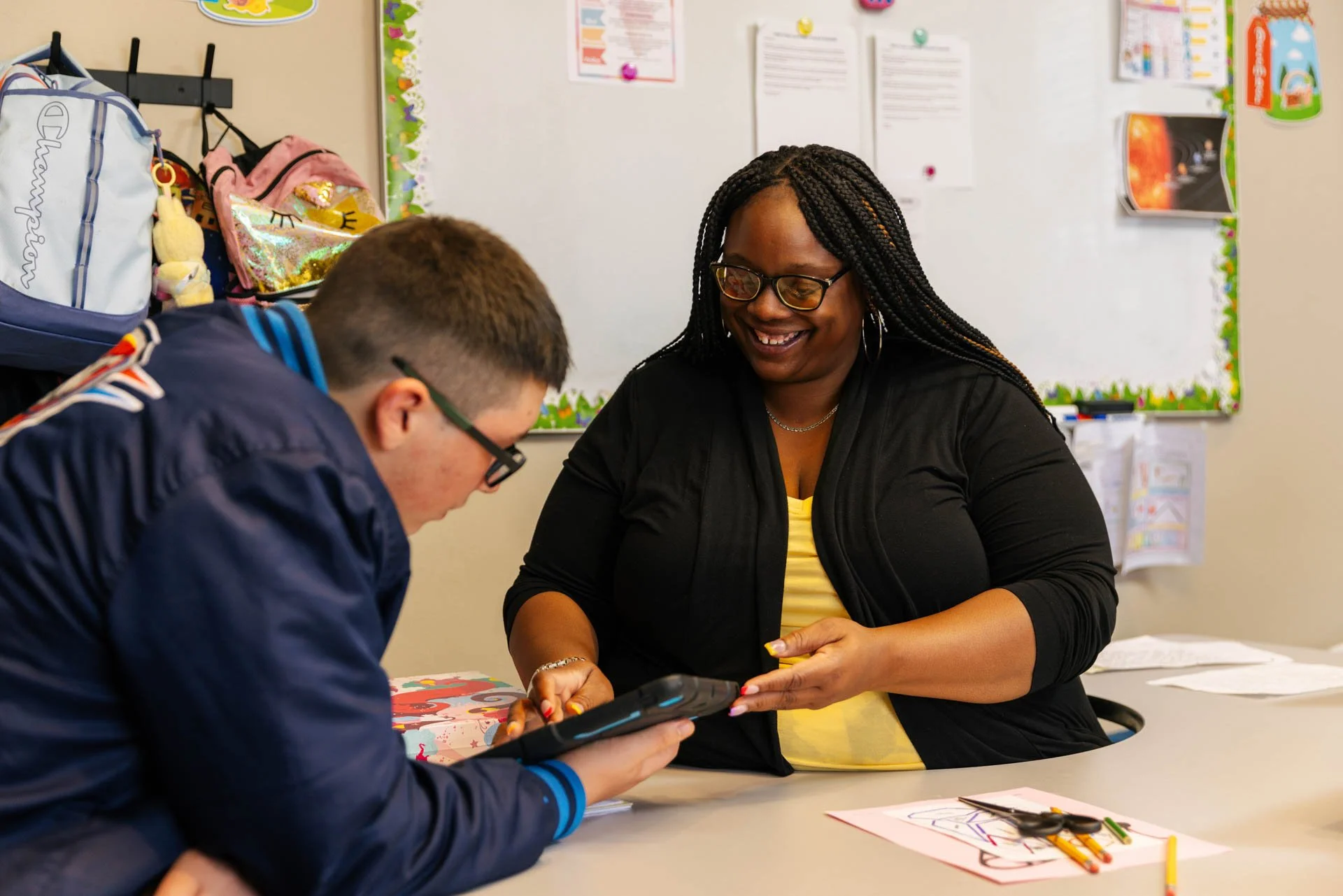 A woman with glasses smiling while talking to a boy at a classroom table. Items such as paper, crayons, and a tablet are on the table. The background features backpacks, a bulletin board with papers and colorful decorations.