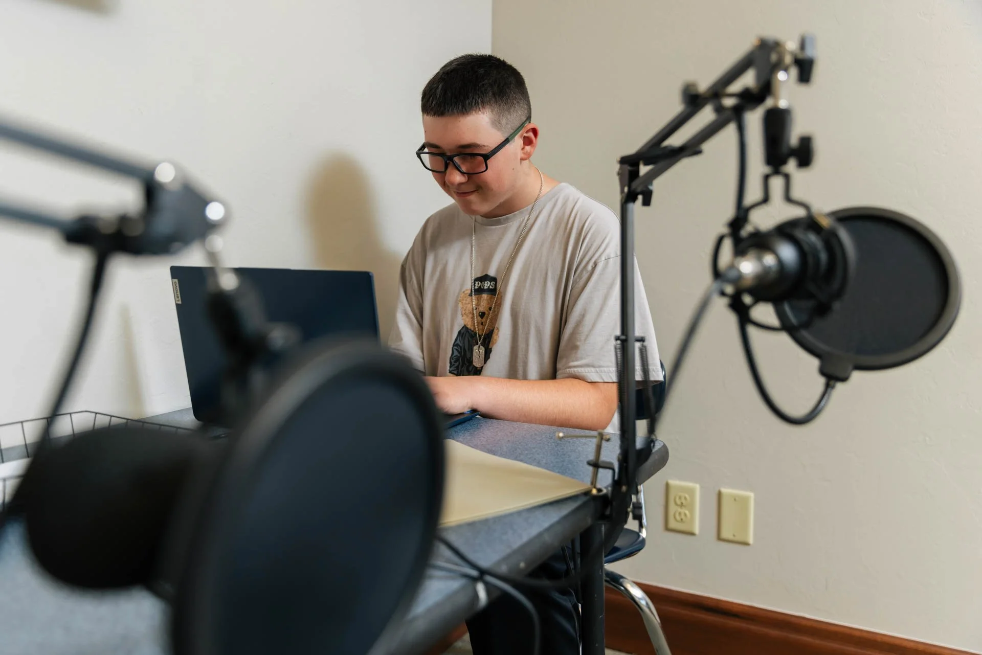 A young man with glasses and short dark hair sitting at a desk, working on a laptop, with professional audio equipment in the foreground.
