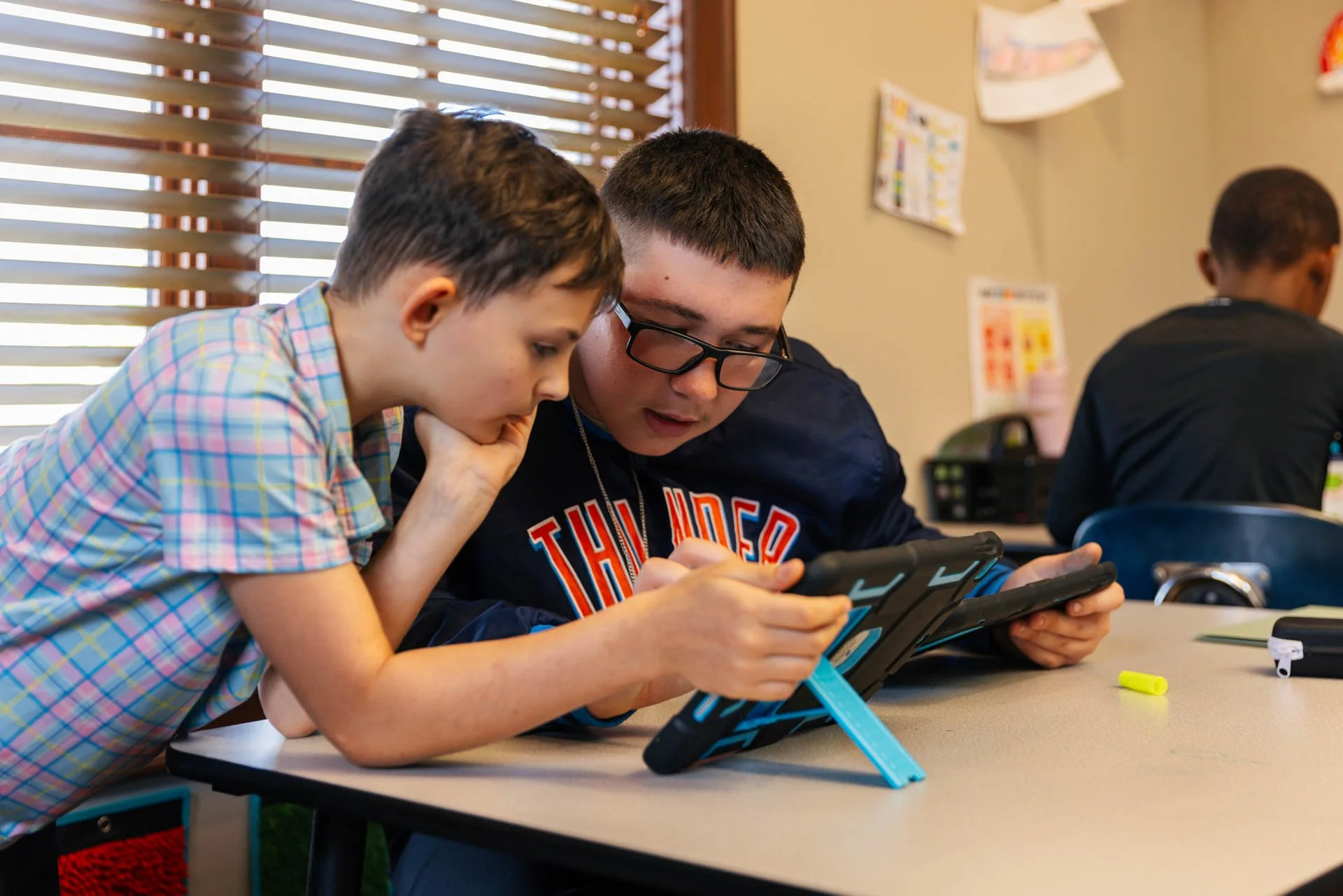Two boys sitting at a table, engrossed in a tablet, with the boy on the left wearing a t-shirt and the boy on the right wearing glasses and a dark sweatshirt, in a classroom with windows and posters on the wall.