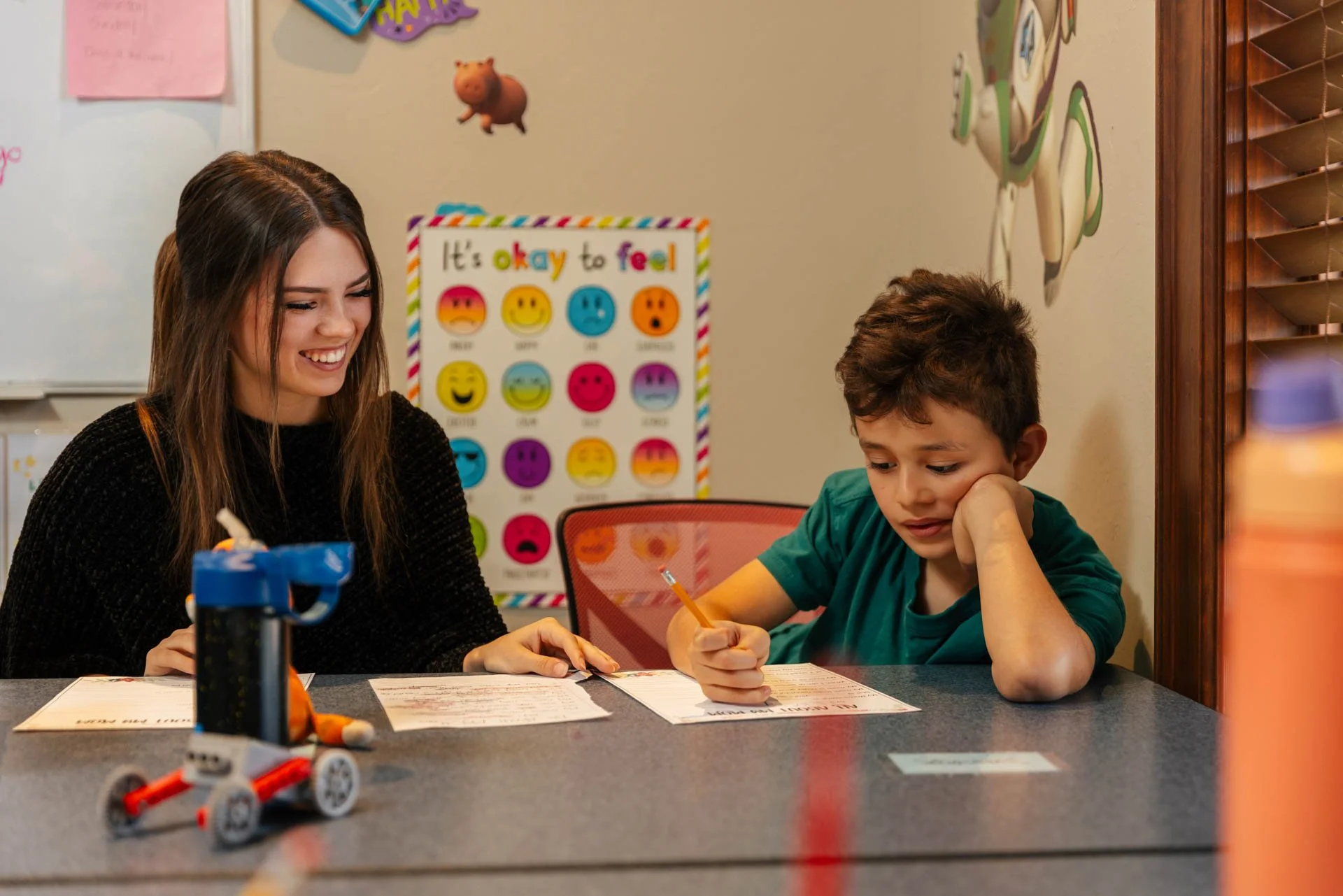 A woman and a young boy sitting at a table in a classroom or study area. The woman is smiling and looking at the boy, who is focused on writing. There is a colorful poster of emojis behind them with the phrase 'It's okay to feel'. A robot toy or model is on the table in the foreground, and a window with wooden blinds is on the right side of the image.