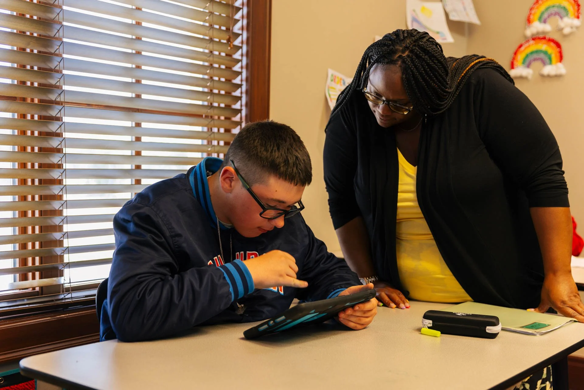A teacher standing next to a student who is sitting at a table, looking at a tablet device. The scene is indoors with window blinds in the background decorated with children's drawings and rainbows.