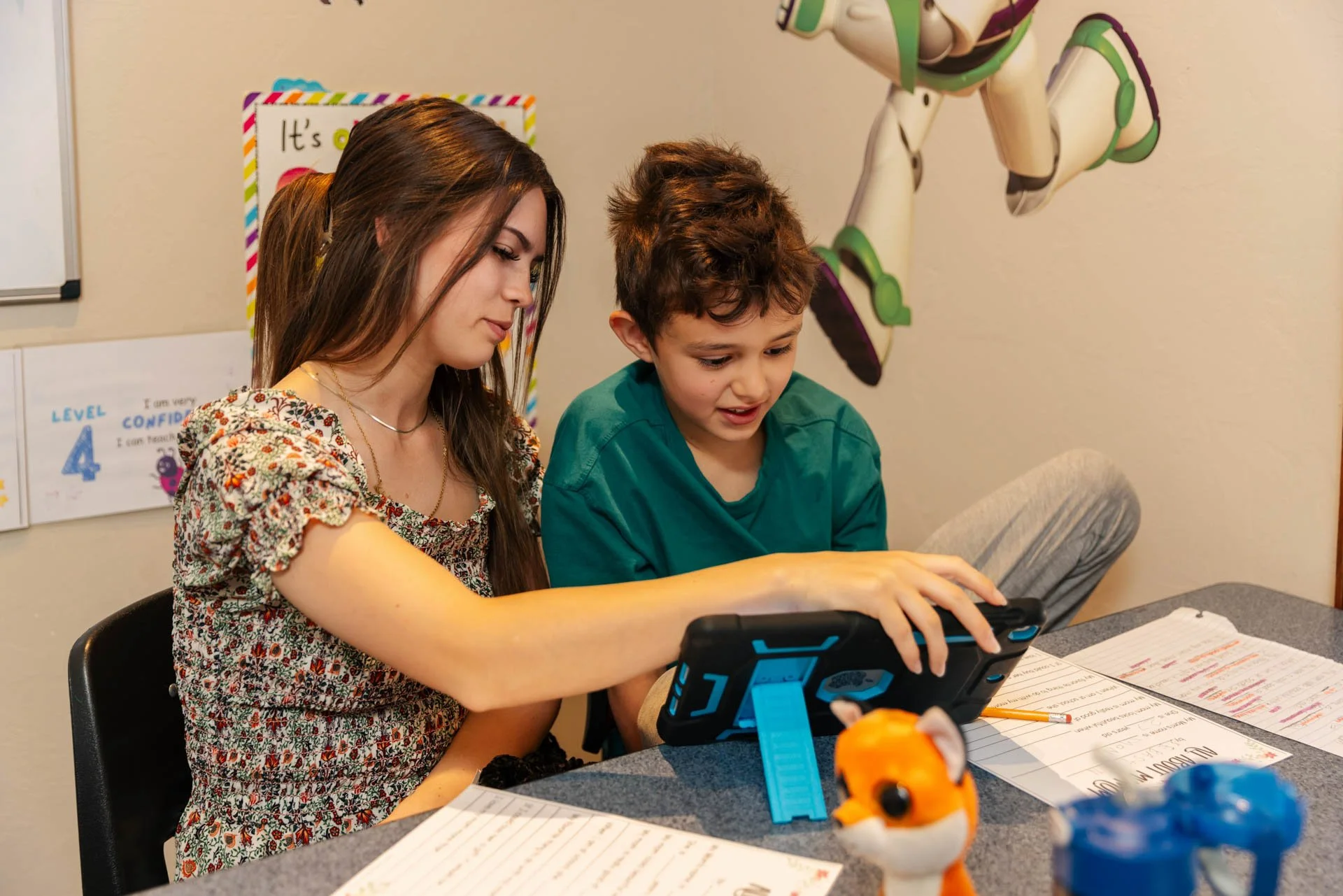 A young woman with long brown hair and a young boy with short brown hair sit at a table in a classroom, looking at a tablet device together. The table has papers, a pencil, a small orange toy, and a toy dog in the foreground. The classroom wall behind them is decorated with colorful posters and a large robot figure.