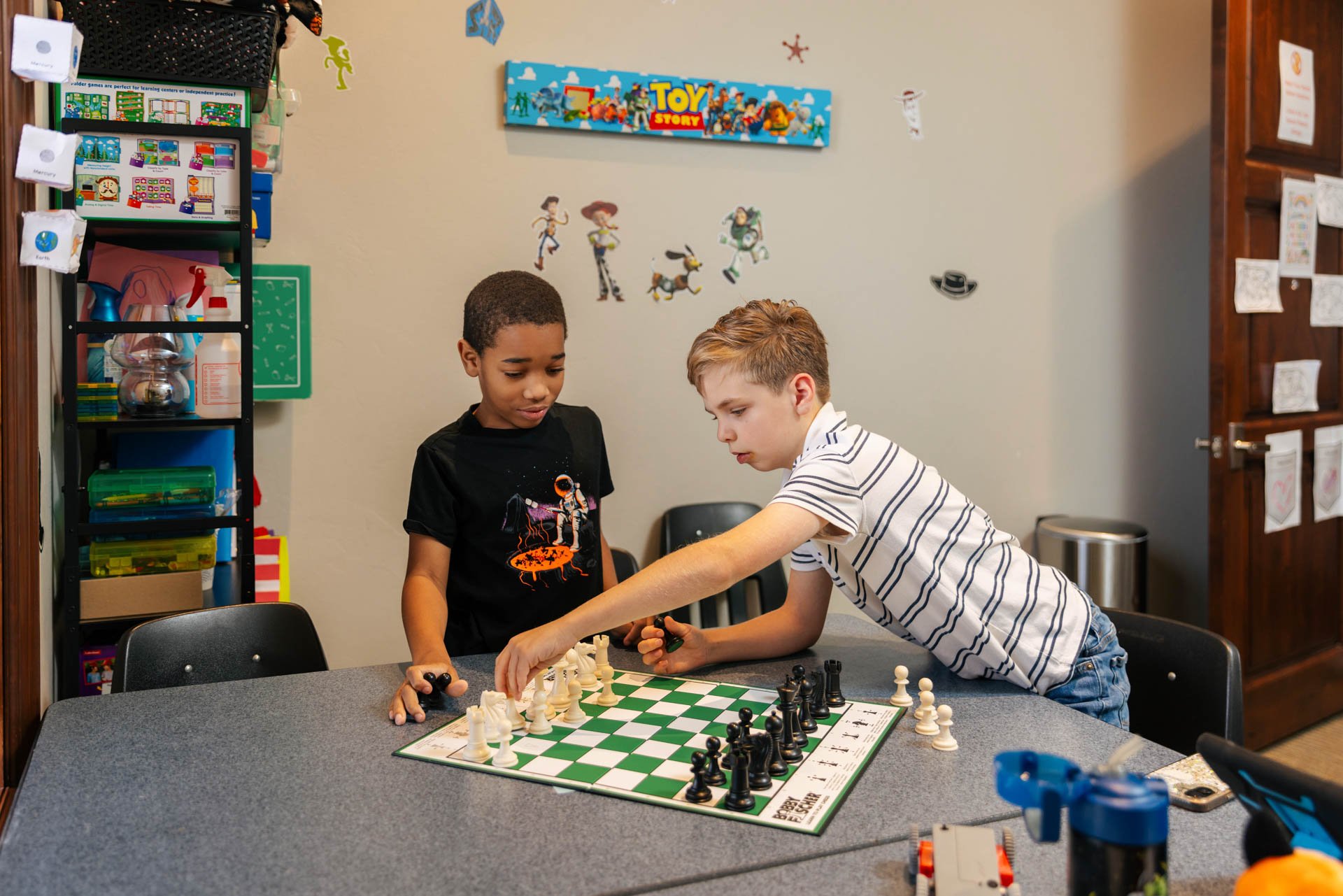 Two boys playing chess at a table in a game room decorated with Toy Story themed wall stickers, including characters and a banner, with shelves containing colorful toys and supplies nearby.