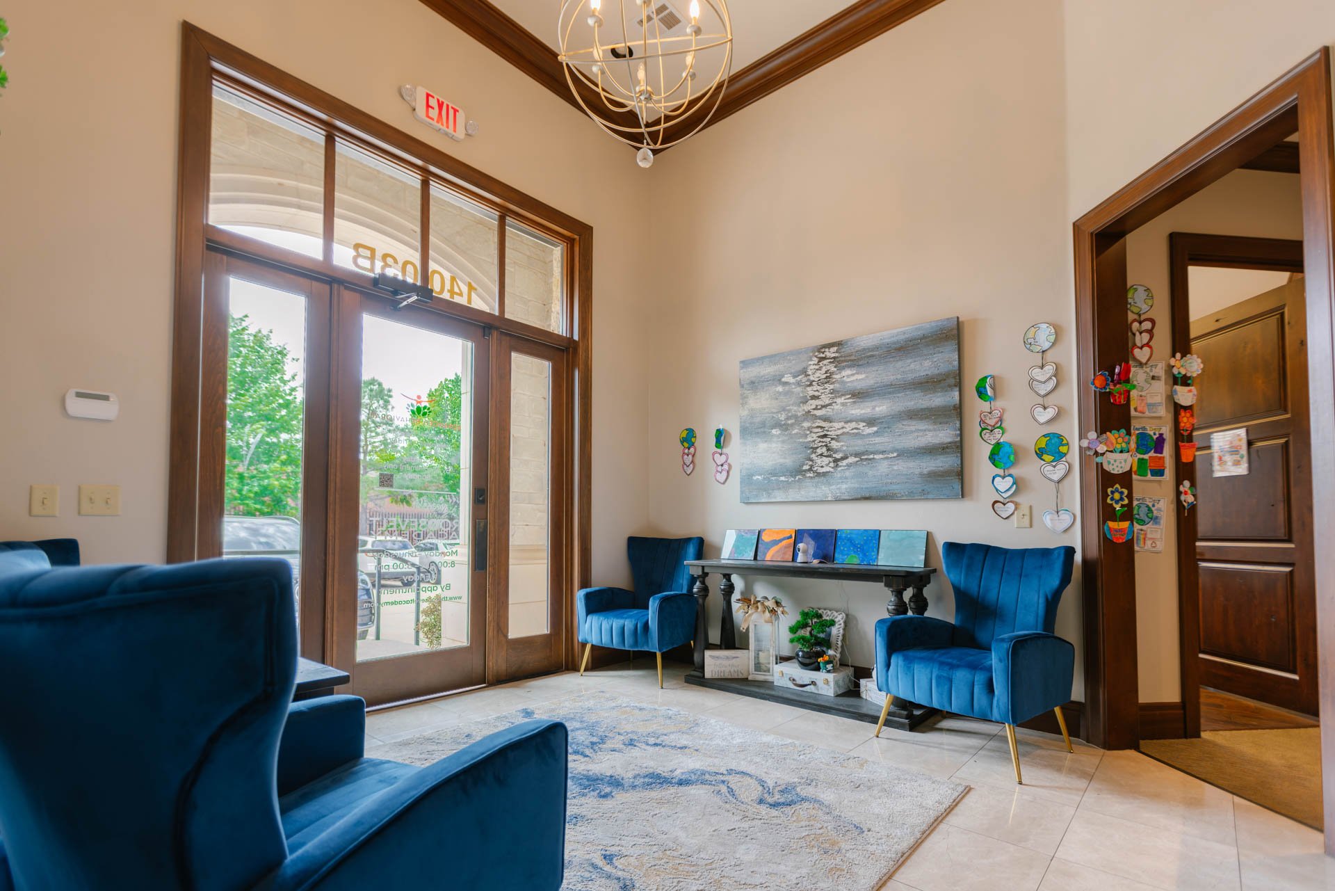 A cozy waiting area with blue velvet chairs, a table with artwork, and colorful hanging decorations on the wall near wooden door and large glass entrance