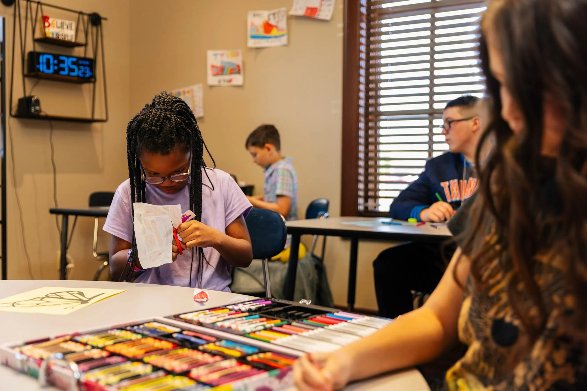 Young girl with braided hair and glasses sitting at a table, opening a gift bag. Several children sit at desks or tables in a classroom with educational posters and a digital clock on the wall.