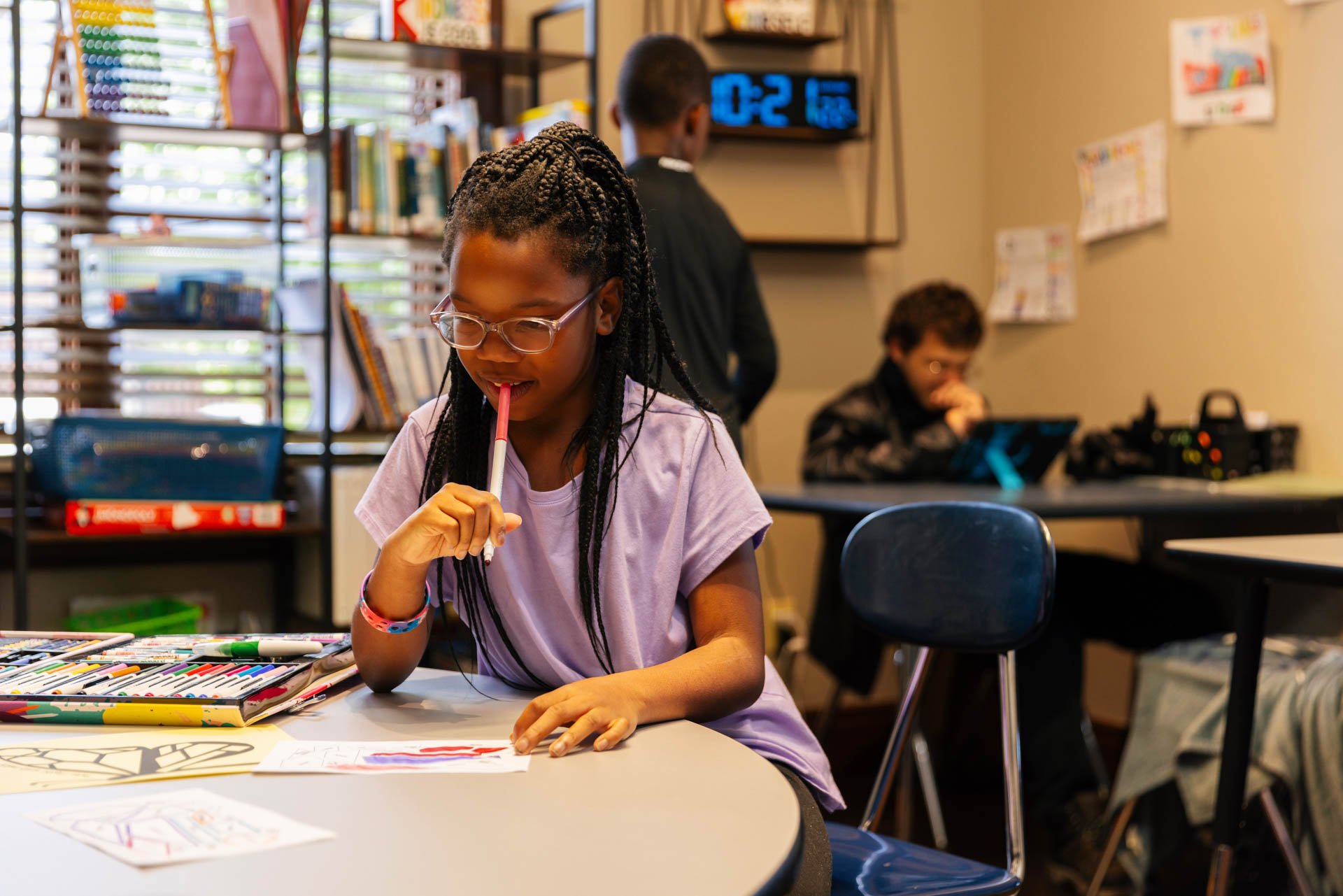 A young girl with glasses and braided hair sitting at a table, holding a colored pen to her mouth, working on an art project in a classroom. In the background, other children are at work.