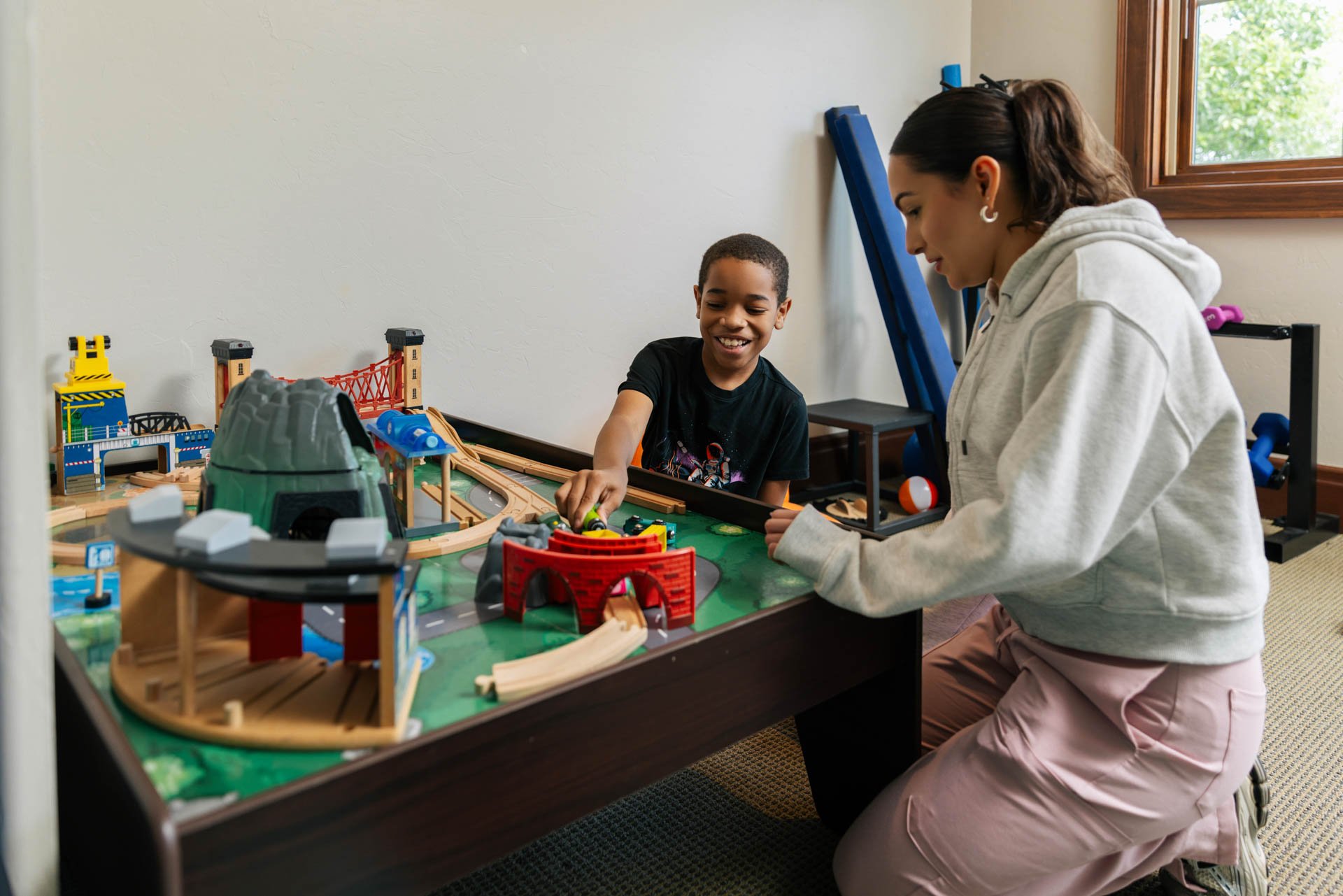Two kids playing with a large train set on a table in a room with a window.