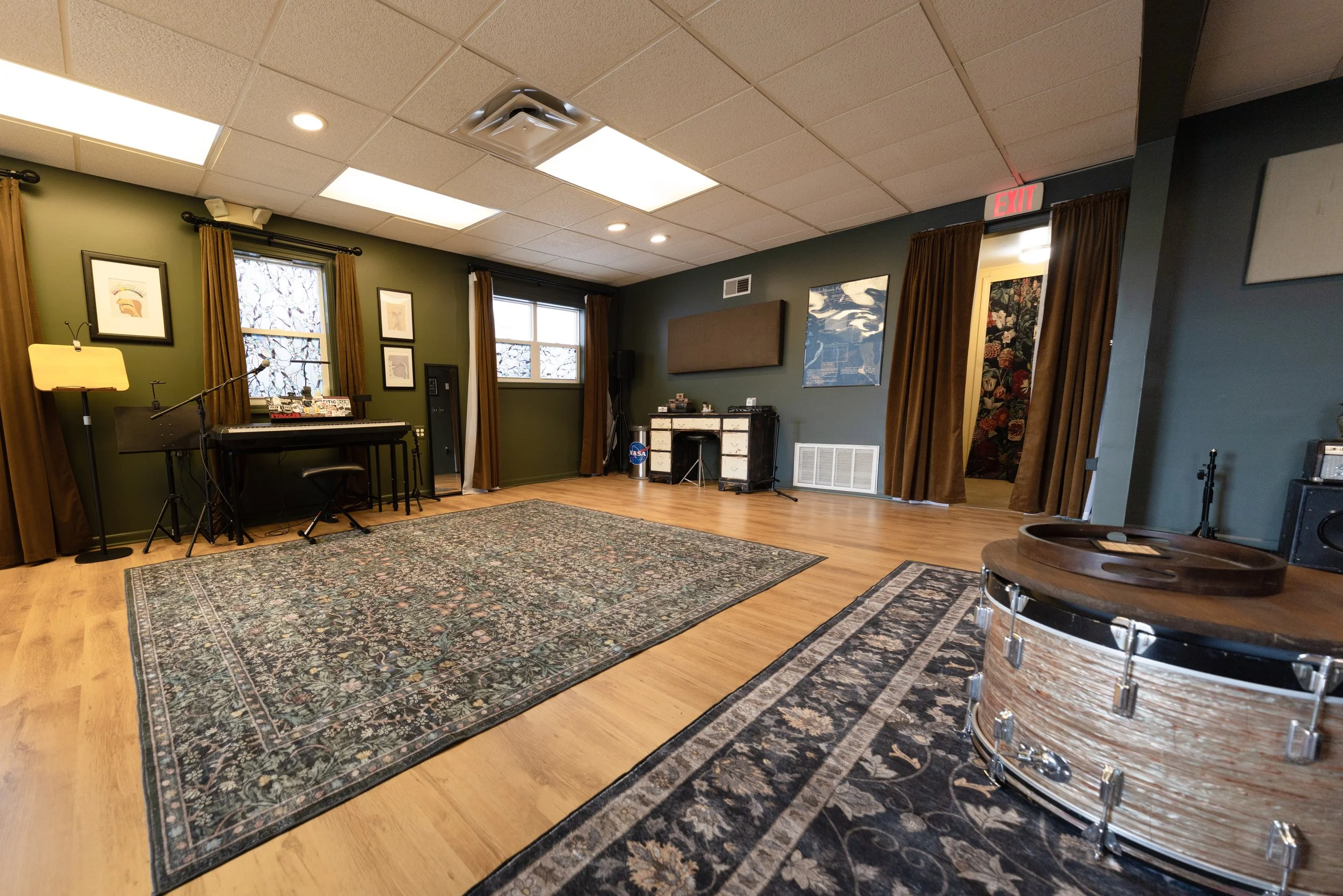 An indoor room set up for a musical performance with a piano on the left, a drum in the foreground on the right, and various framed pictures on the green walls. There are windows with brown curtains, and a rug on a hardwood floor.