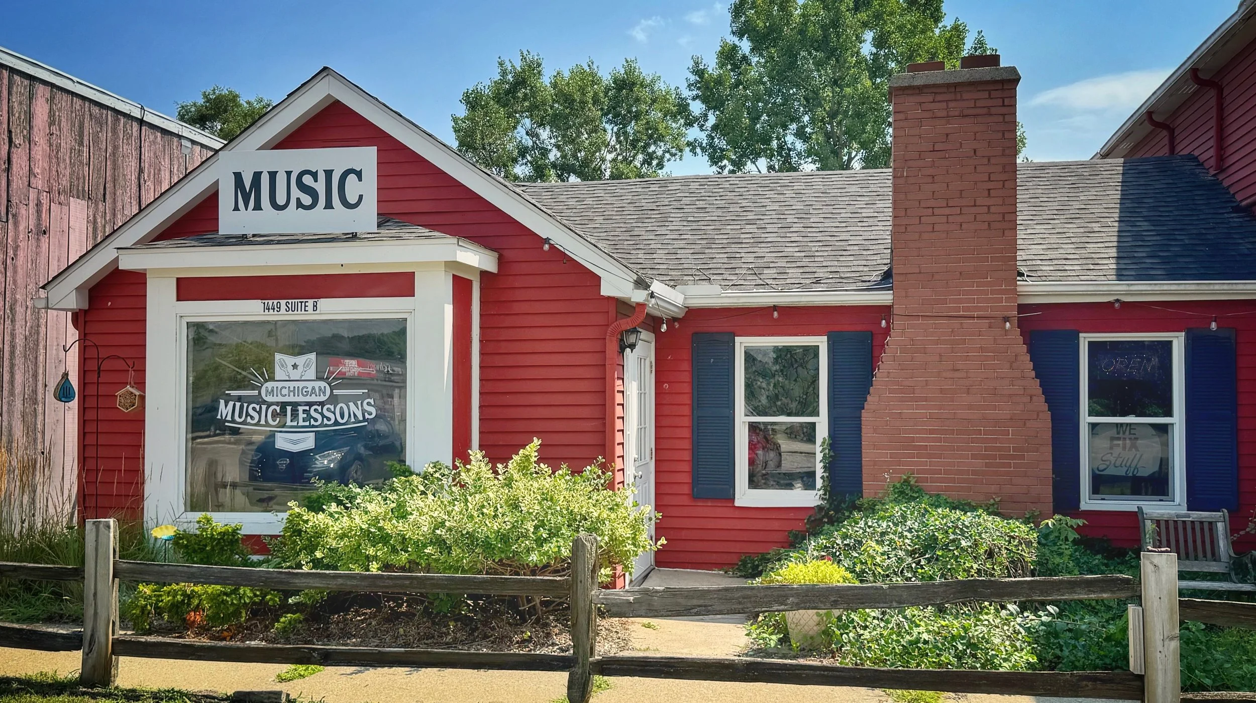 Red building with a large sign that reads 'MUSIC', a window with a sign that says 'Michigan Music Lessons', and an open sign in the window. There are blue shutters and a red brick chimney. Green bushes and a small wooden fence are in front of the bui