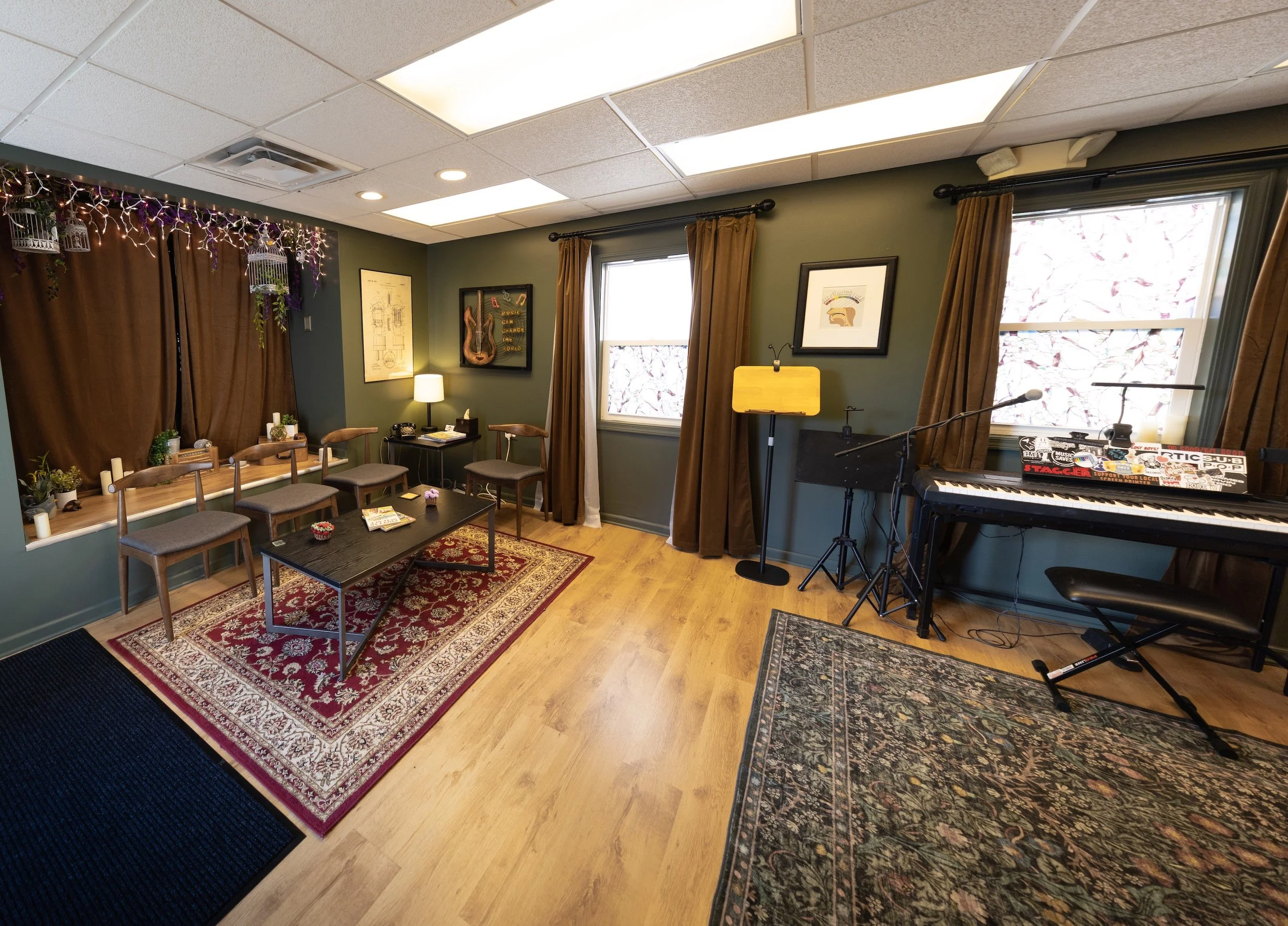 A cozy room with green walls and hardwood floors, decorated with framed artwork, a small table with a lamp, and a row of wooden chairs, featuring a corner with a keyboard and microphone, and accented with curtains, rugs, and string lights.
