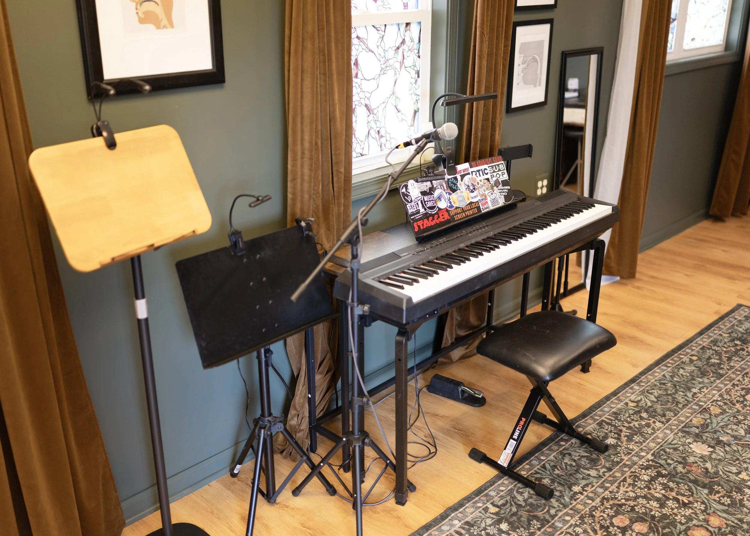 A music practice space with a digital keyboard on a stand, a music stand, a chair, a microphone on a stand, and framed artwork on a green wall with a window and brown curtains.
