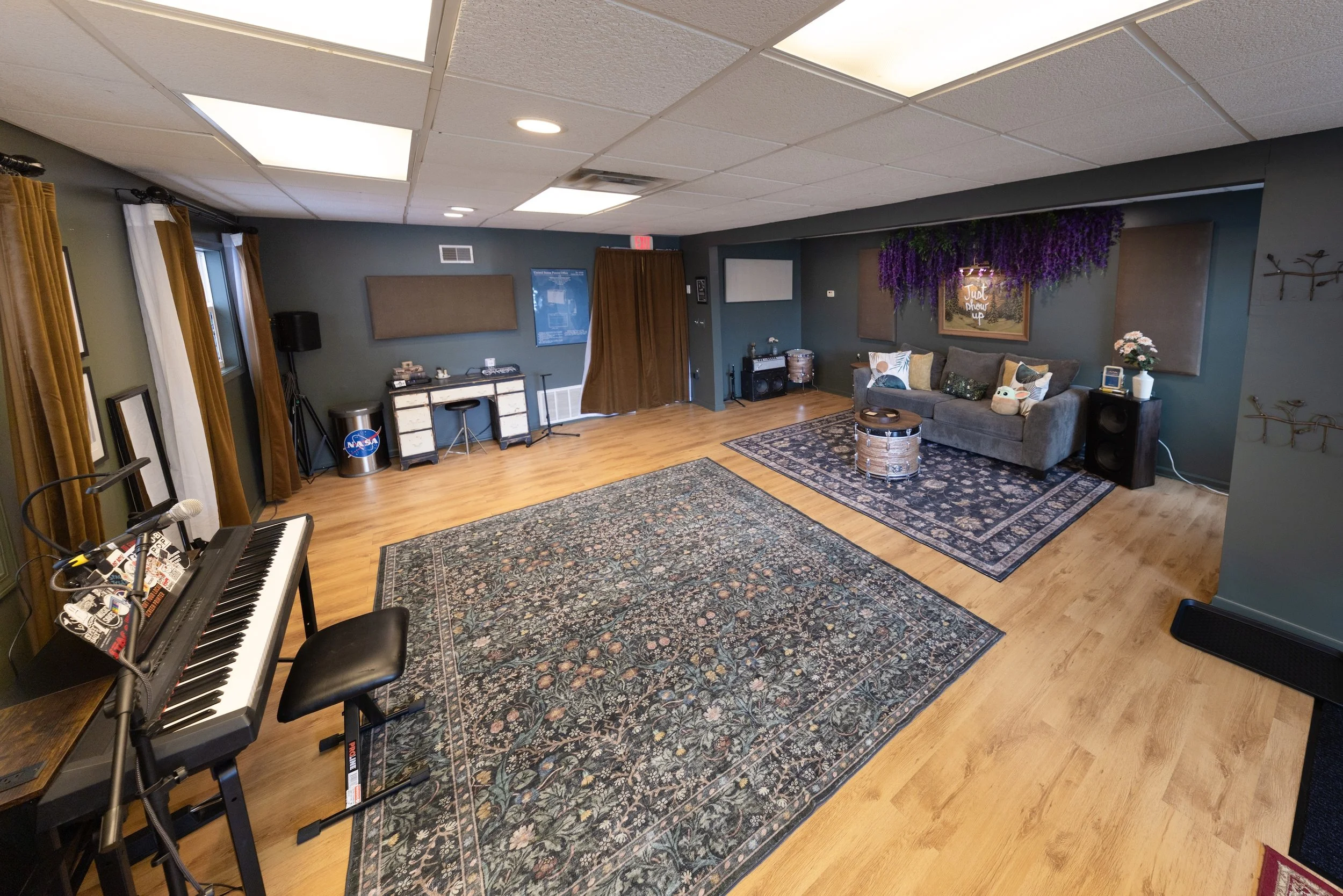 A spacious room with wooden flooring, dark teal walls, and a white drop ceiling with fluorescent lights. It features a black keyboard on a stand with a stool, a pair of curtains beside windows, a gray sofa with decorative pillows, a vintage drum, a p