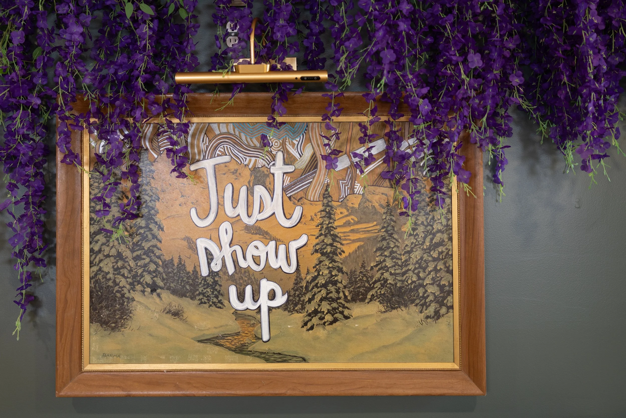 A framed wall decoration with a mountain landscape painting and the words 'Just breathe up' in white text. Purple hanging flowers hang over the frame, and a gold-colored device is mounted on top of the frame.