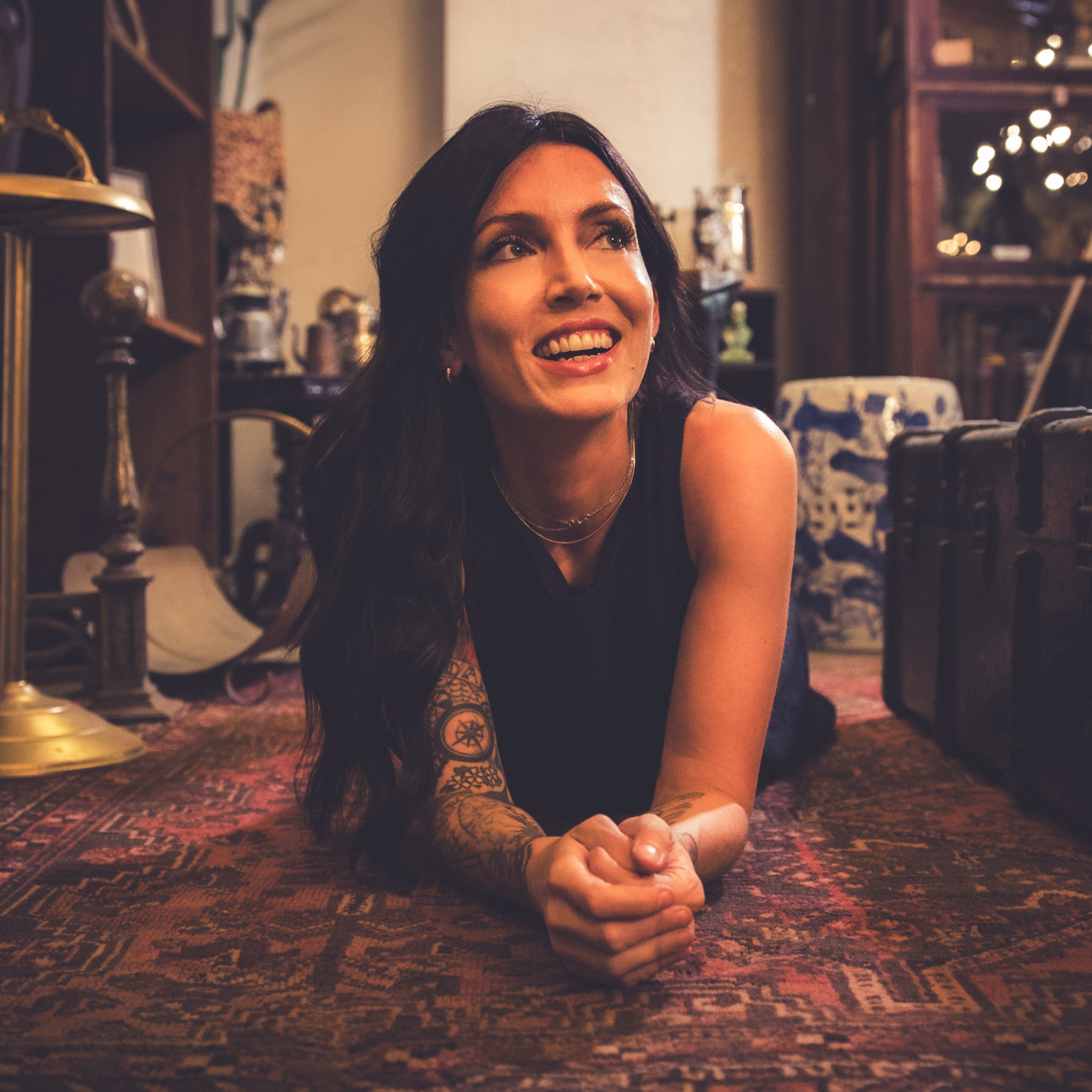 A woman with dark hair and tattoos on her arm smiling and looking upward while lying on her stomach on a patterned rug indoors.