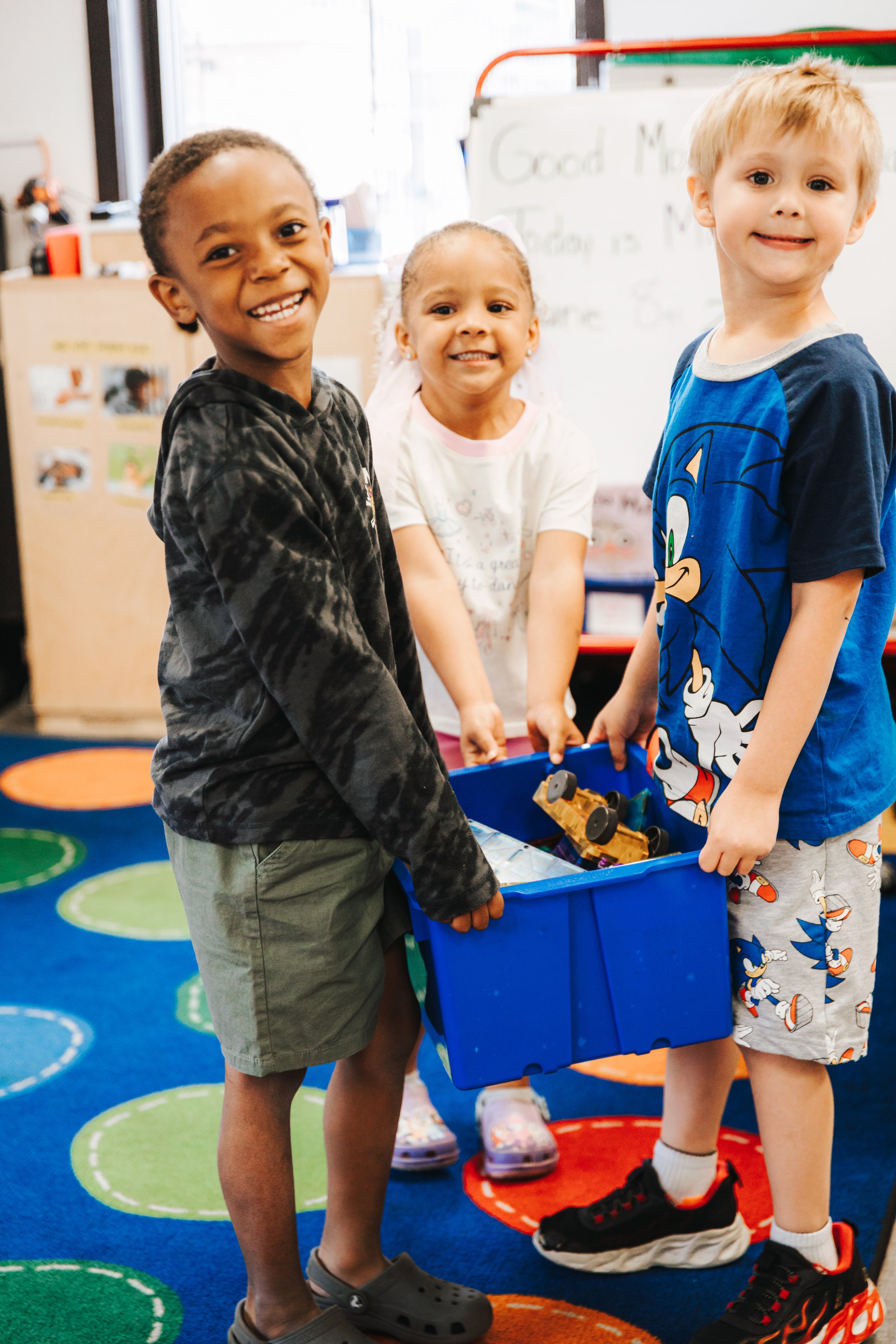 Three children standing in a classroom, holding a blue plastic container filled with toy cars, smiling at the camera. The background includes a whiteboard and colorful circular carpet patterns.