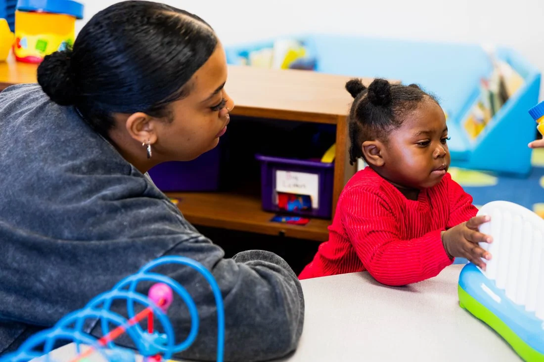 An adult and a young girl with braided hair sitting at a table in a classroom, engaging in a learning activity with educational toys.
