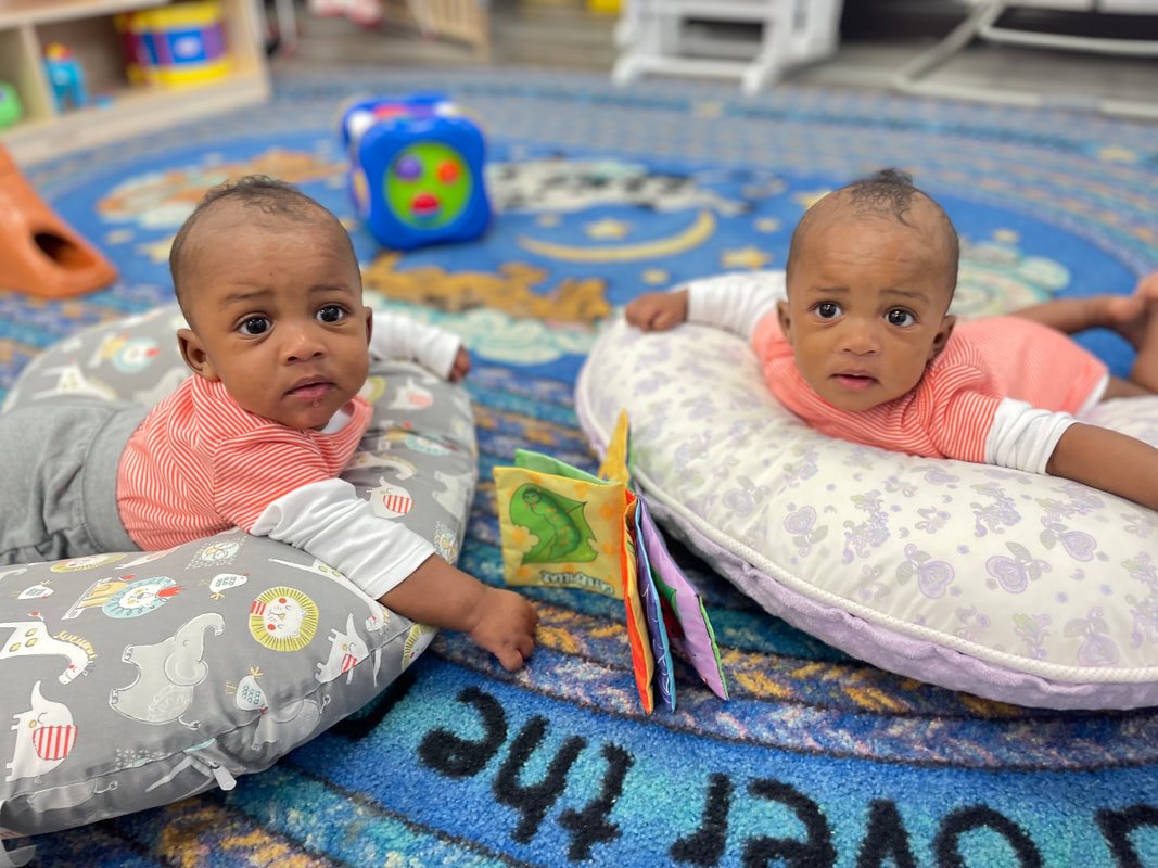 Two babies lying on their stomachs on colorful mats, looking at the camera, with toys and blankets around them.