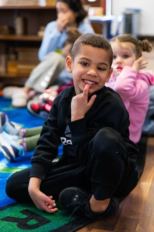 A young boy sitting on a colorful rug smiling and touching his chin with his finger, with two girls and an adult blurred in the background.
