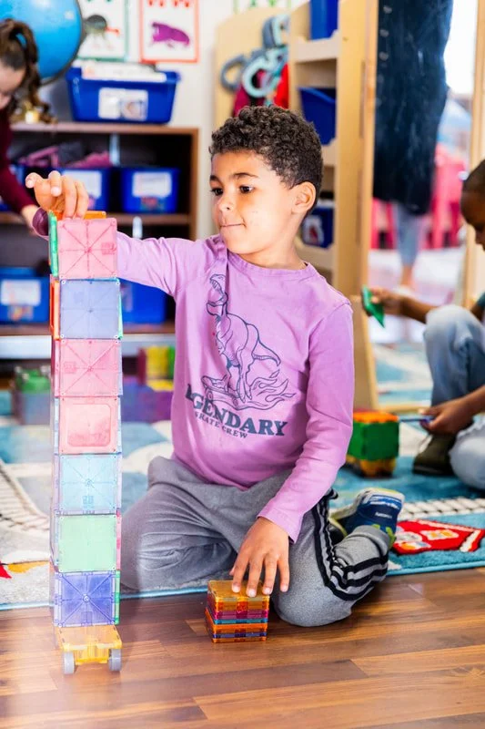 A young boy in a pink long-sleeve shirt stacking colorful plastic tiles into a tall tower in a classroom or playroom.