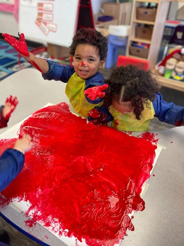 Children playing with red paint while making art at a table in a classroom.