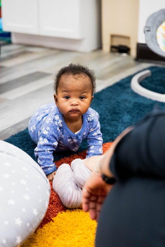 A baby girl with curly hair, wearing a blue star-patterned pajama, is crawling on a multicolored rug in a living room, looking at an adult's hand.