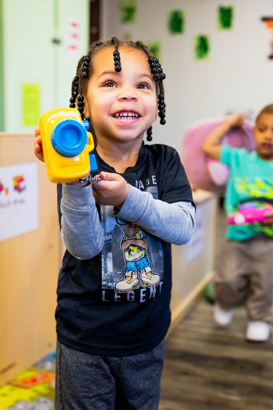 A young girl with braided hair holding a yellow toy camera, smiling. In the background, another child is leaning against a wall, holding a pillow.