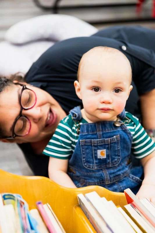 A woman with glasses and dark hair leaning over a young child with a bald head, sitting in front of a yellow bookshelf with colorful books, in an indoor setting.