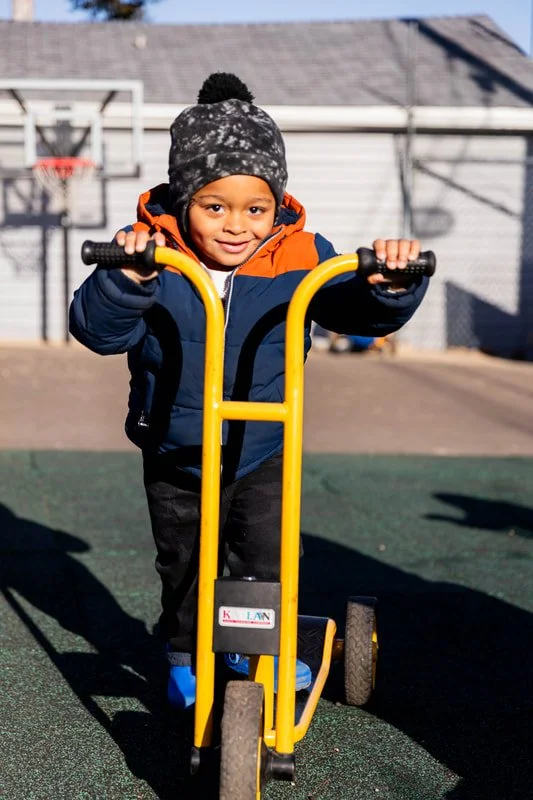 Young child wearing a jacket and hat, riding a yellow scooter at a playground during daytime.