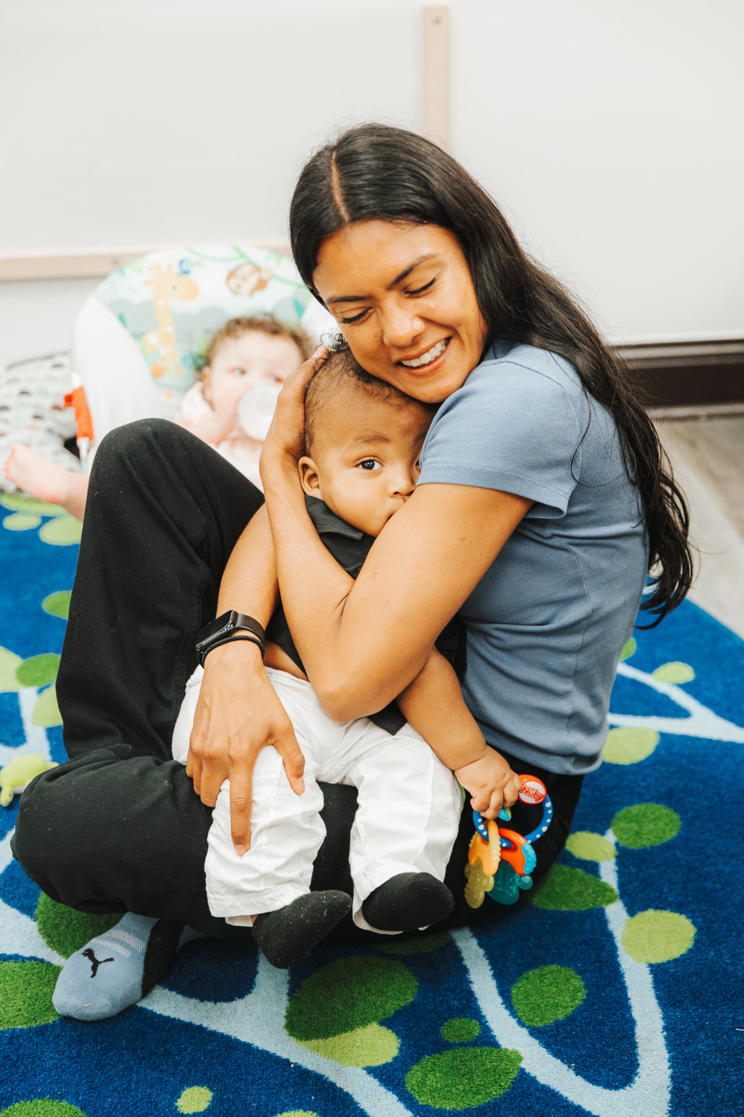 A woman with long black hair hugging a young boy with short black hair, who is holding a colorful toy. In the background, a girl with a pacifier in her mouth is sitting in a padded seat on a colorful blue and green patterned carpet, with white walls and a whiteboard behind them.