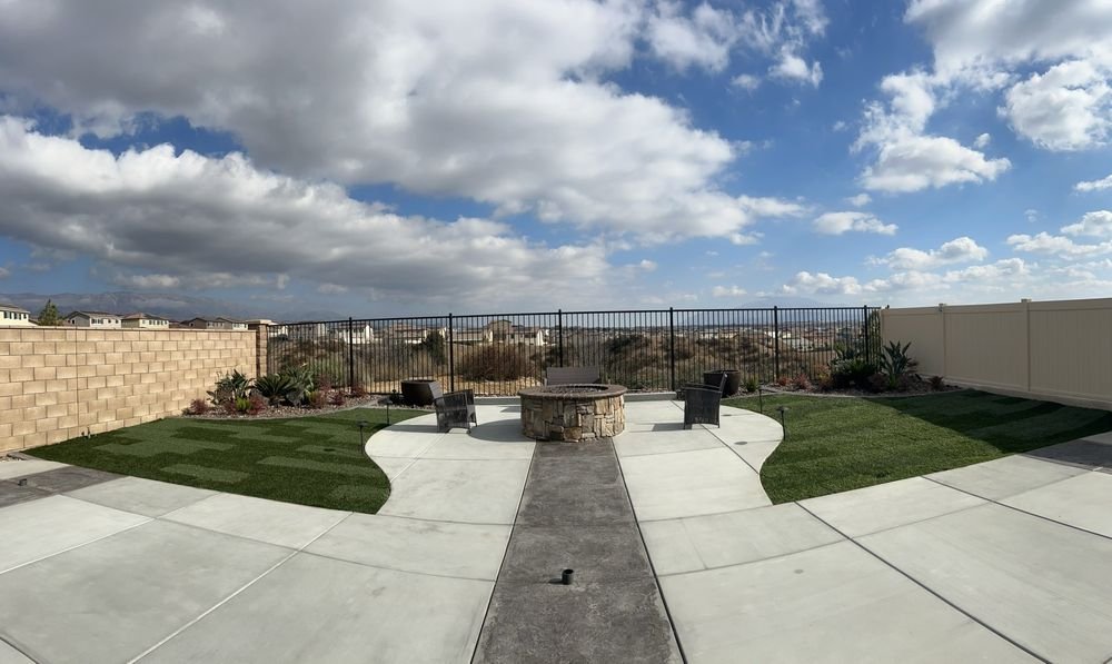 Backyard patio with a fire pit, two chairs, decorative plants, a patio wall, and a view of a suburban neighborhood under partly cloudy skies.