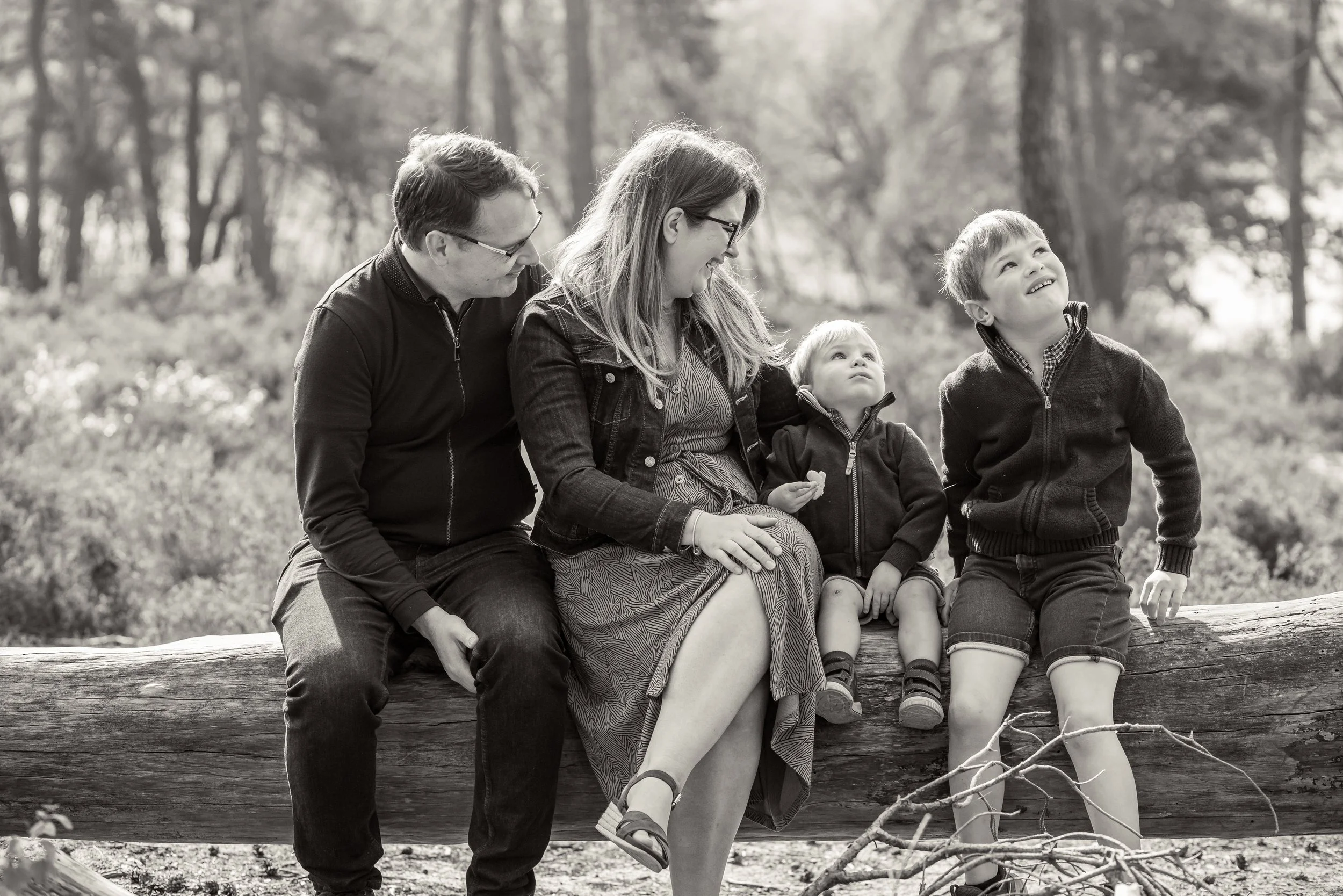 A black and white photograph of a family of five sitting on a log in a wooded area. The family includes a man, a woman, and three children. The man and woman are sitting close together, with the woman smiling while looking at the young boy on her right. The boy is smiling and looking upwards, the youngest girl is sitting next to the woman with a neutral expression, and the middle child, a girl, is sitting with her hands on her lap, looking slightly upward. The background shows trees and a natural outdoor setting.