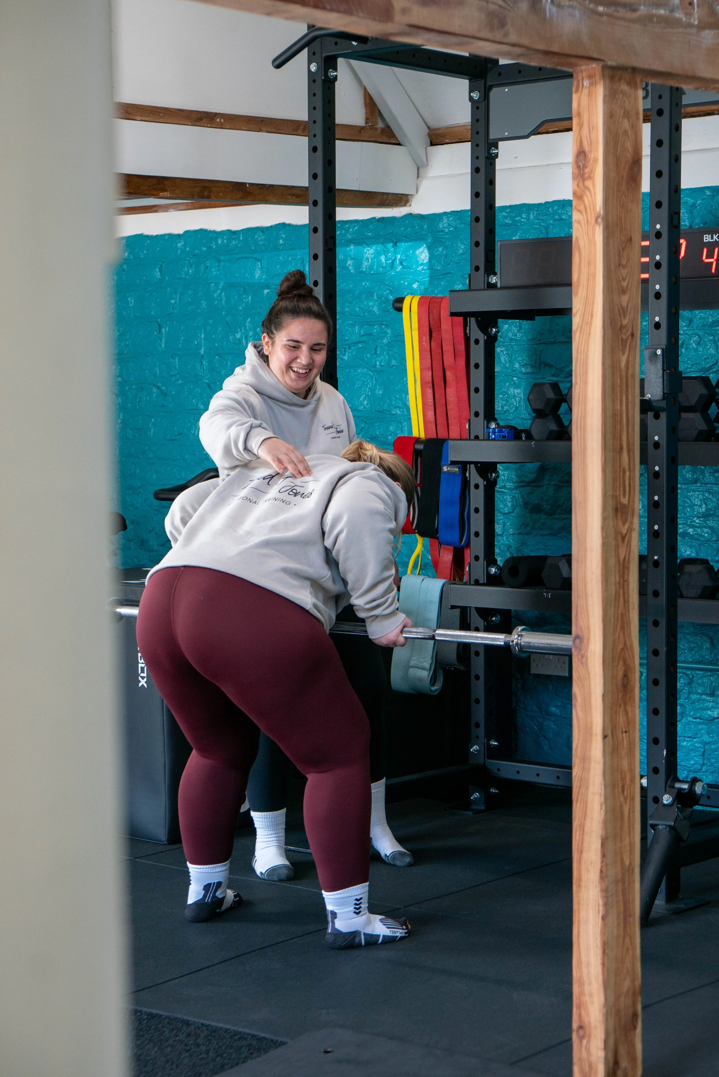 Two women are working together in a gym, with one woman helping the other with a barbell exercise. The setting includes weightlifting equipment, colorful resistance bands, and a textured teal wall in the background.