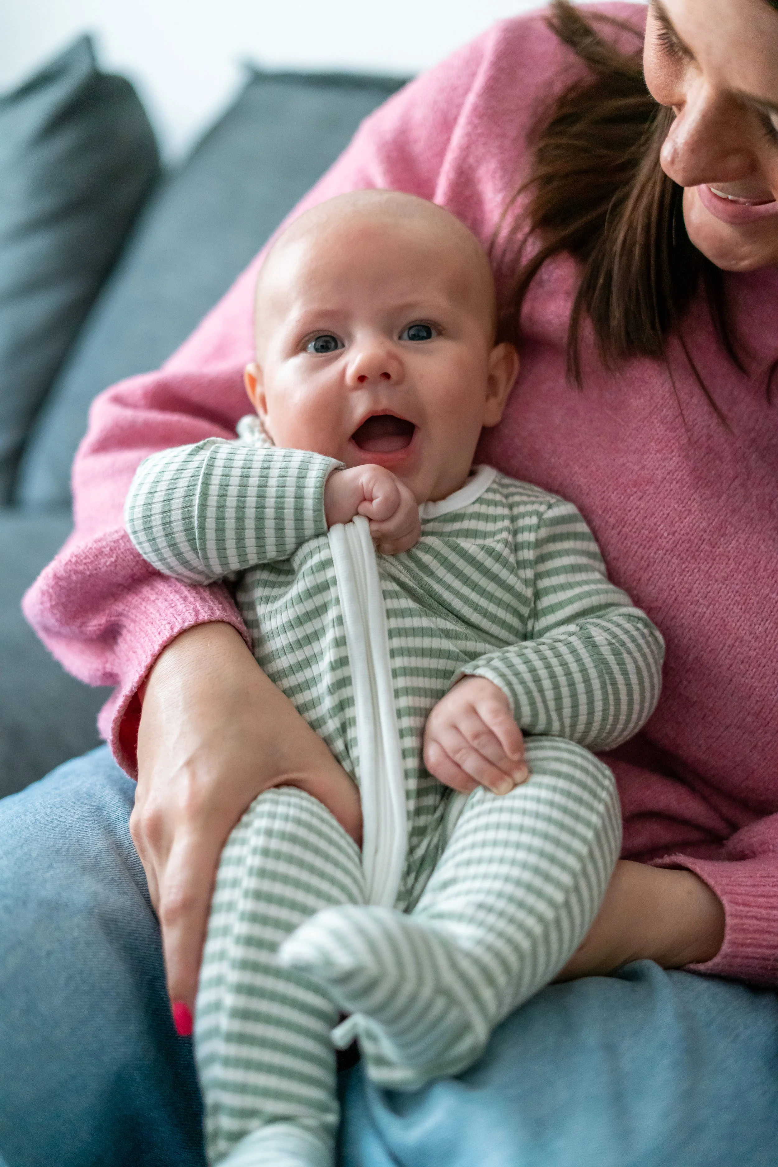 A woman holding a baby on her lap, both sitting on a couch. The baby has wide eyes and an open mouth, wearing a green and white striped onesie. The woman is smiling and wearing a pink sweater.