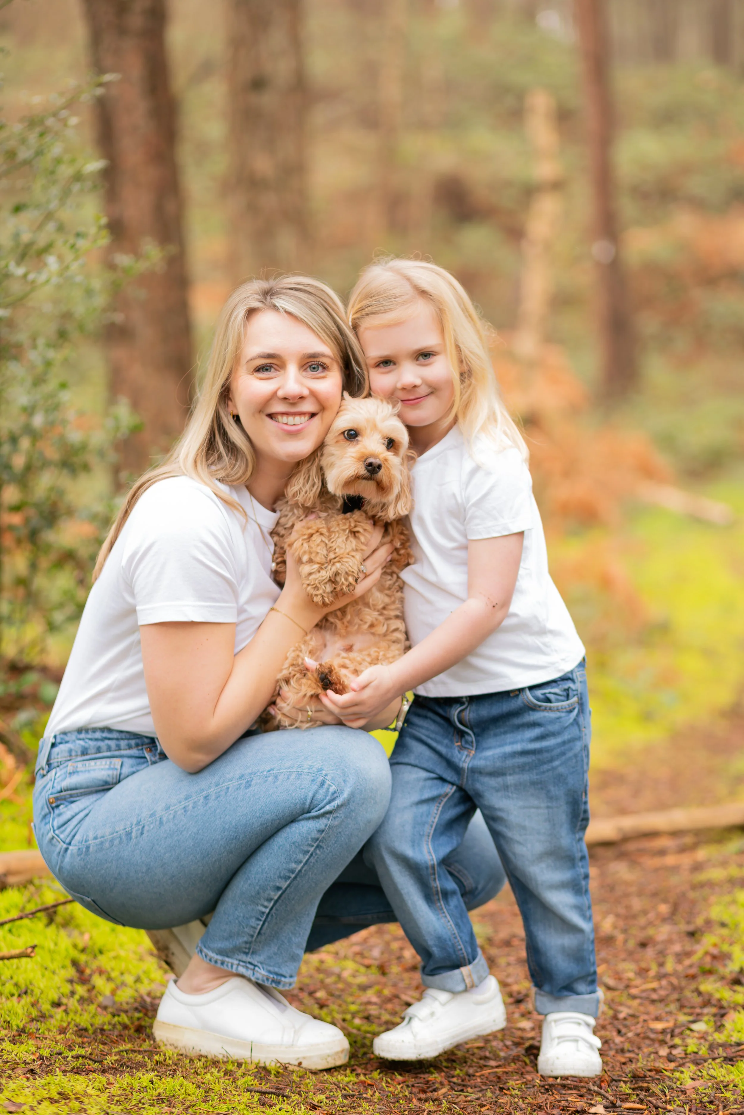A woman and a young girl holding a small dog in a forest with autumn foliage. Both are smiling and dressed casually in white shirts and jeans. Hogmoor Inclosure, Hampshire.