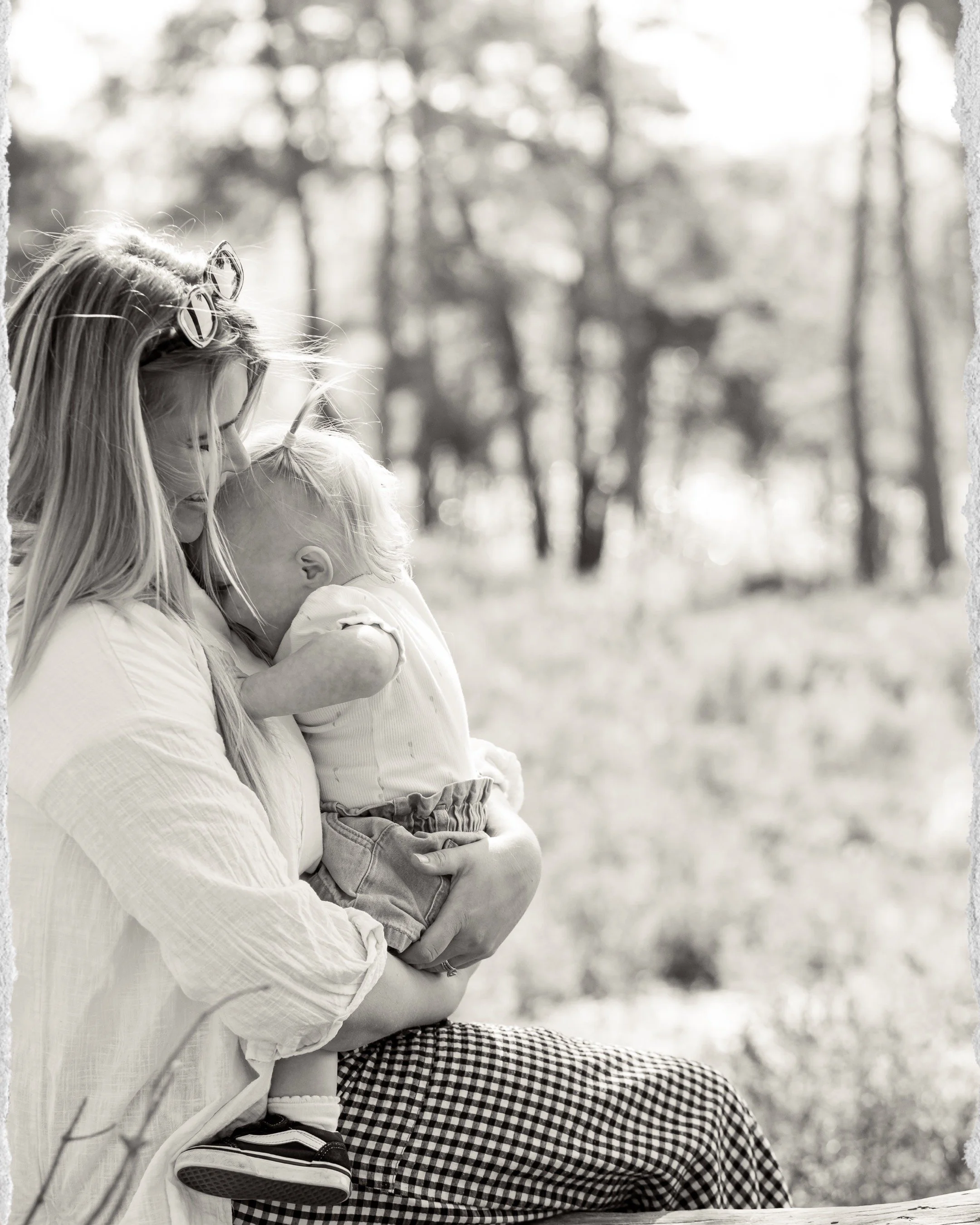 A woman with long hair and glasses holding a young child with short hair in her arms outdoors in a blurred forest setting - Frensham Little Pond, Surrey.