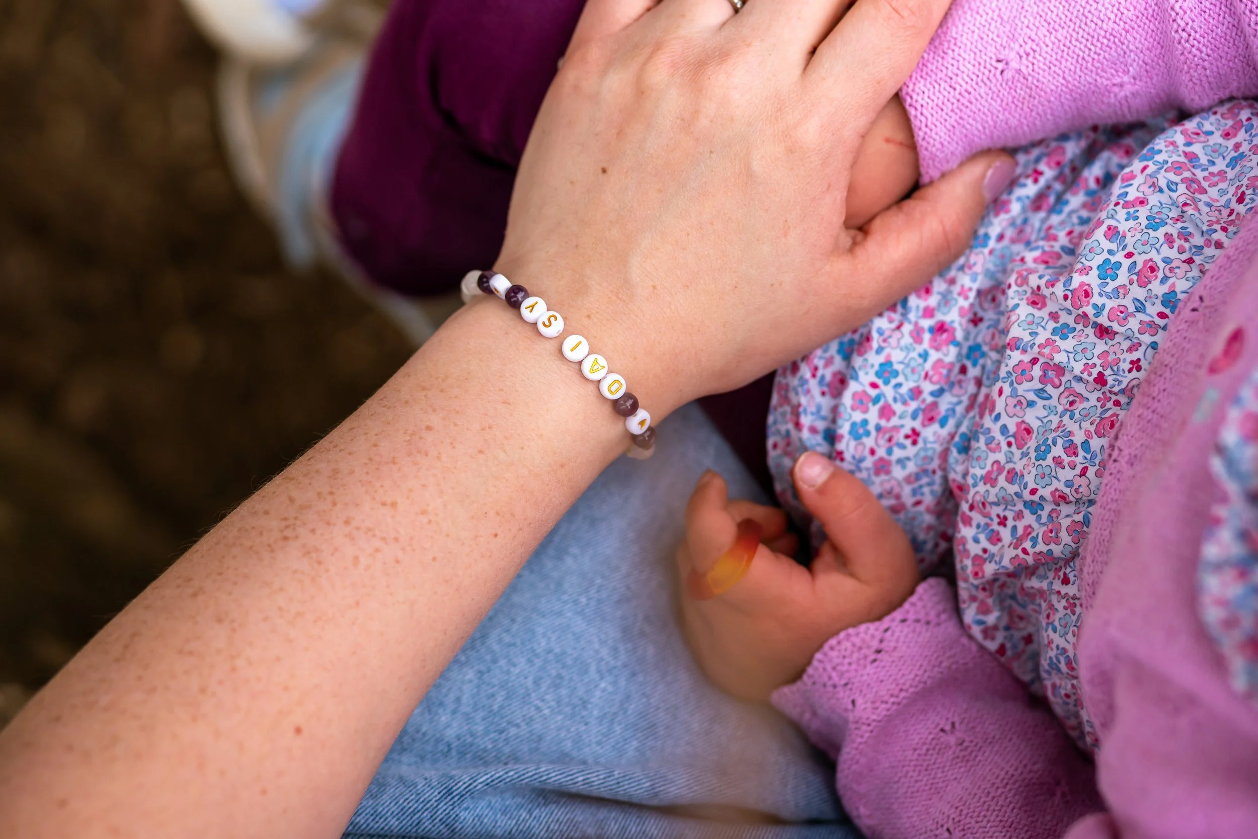 A person's arm resting on a child's chest, with the person wearing a bracelet with white beads spelling out 'DASIA' and dark purple beads. The child is wearing a purple floral dress and pink cardigan, and is holding a small object in their hand.