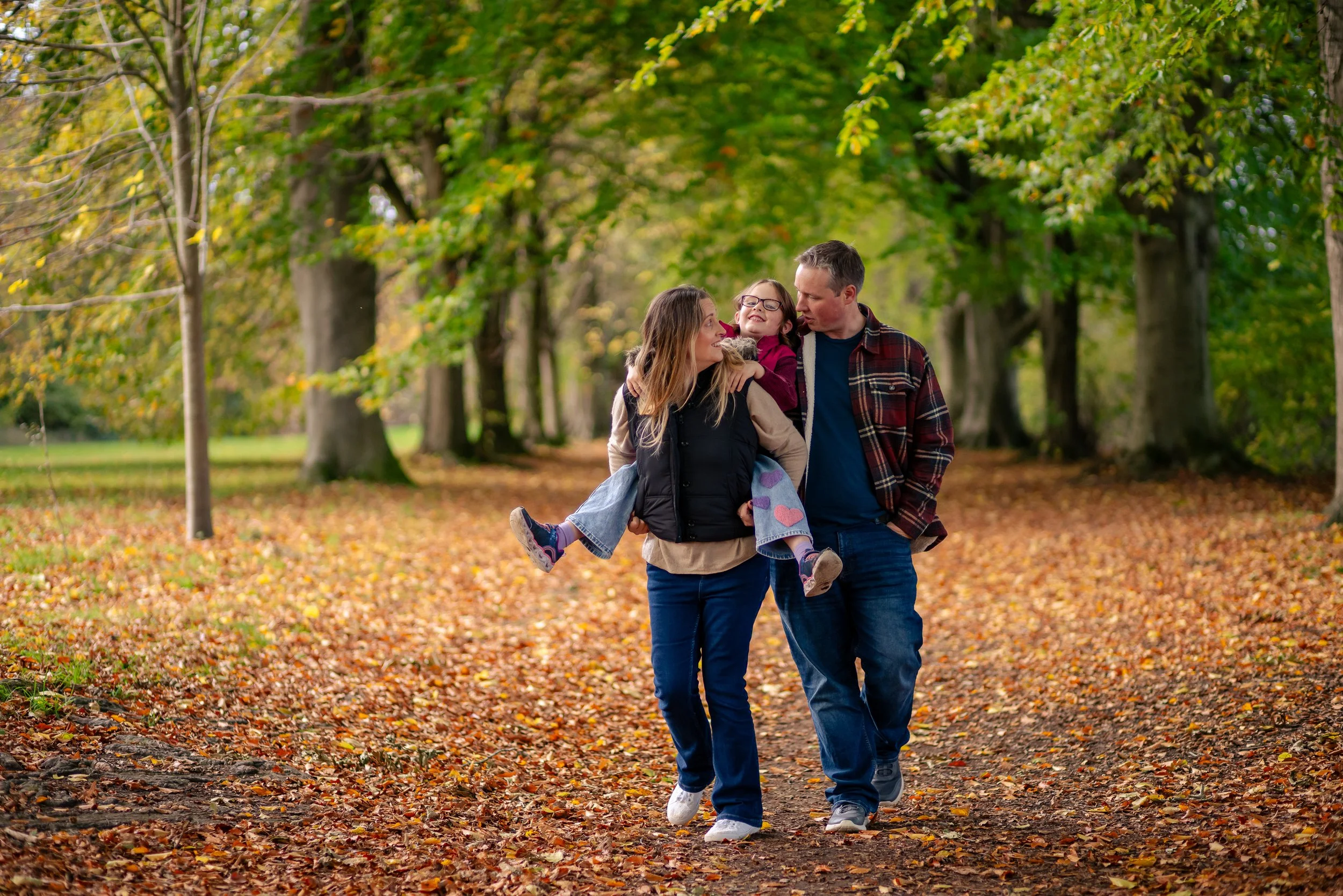 A family of three walking through a park with fall foliage, the mother and father carrying their young daughter on a leash, surrounded by colorful autumn leaves. Farnham Park, Surrey