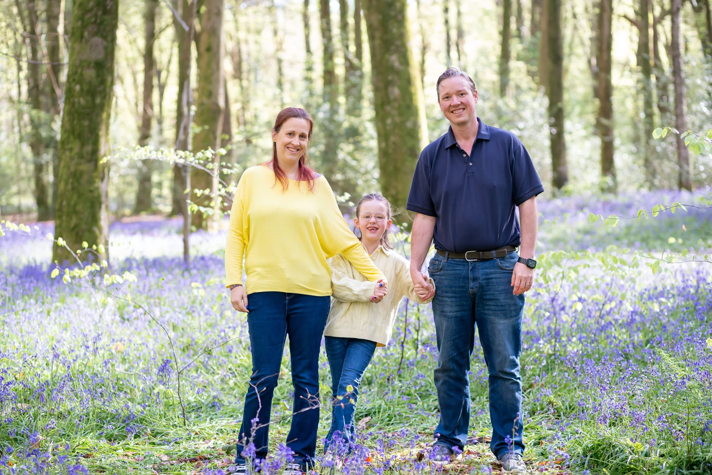 A happy family consisting of a woman, a man, and a young girl walking through a forested area with purple flowers on the ground. They are smiling and holding hands - Micheldever Woods, Hampshire