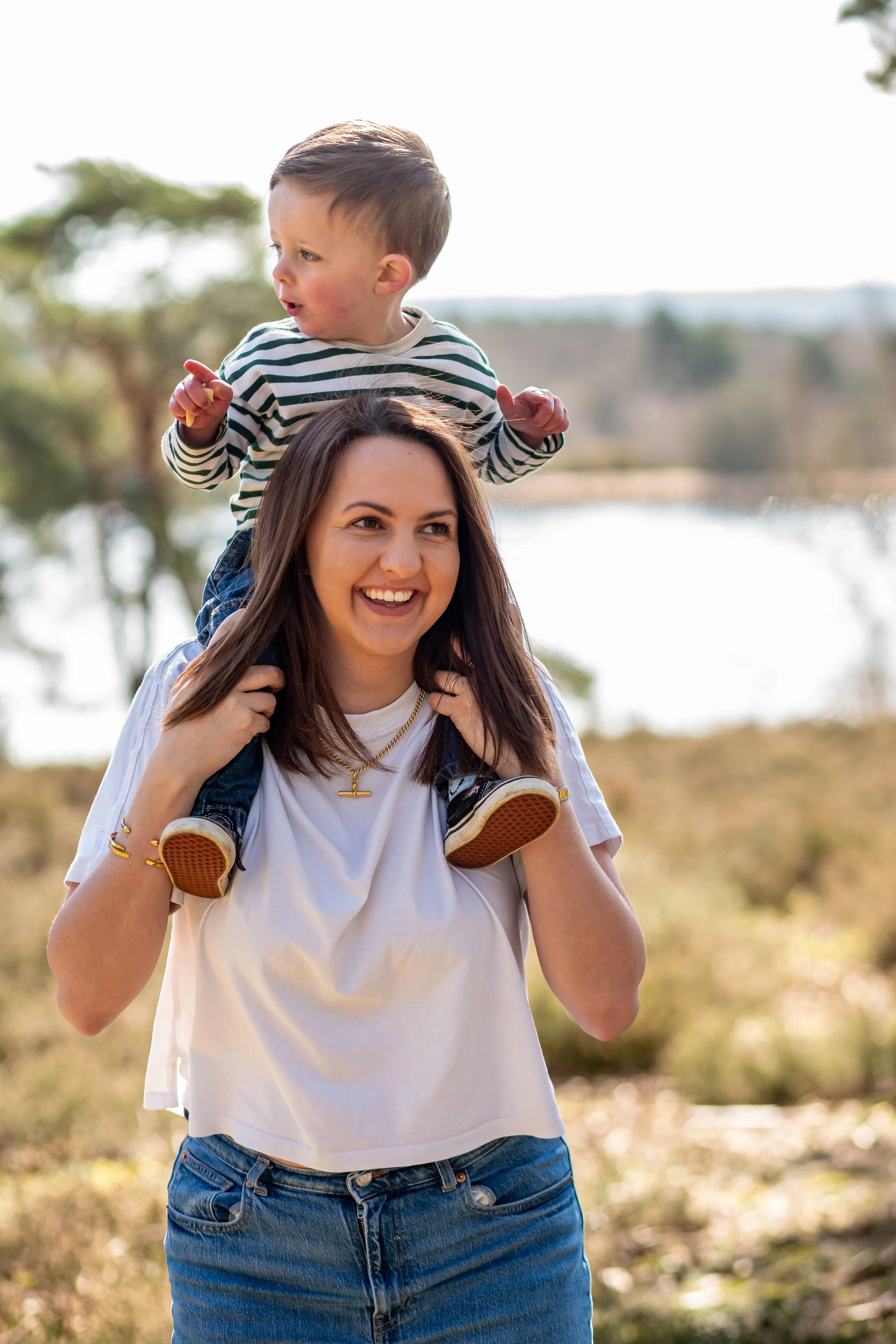 A woman carrying a young boy on her shoulders outdoors near a body of water, with trees in the background, in a sunny setting.- Frensham Little Pond, Surrey.