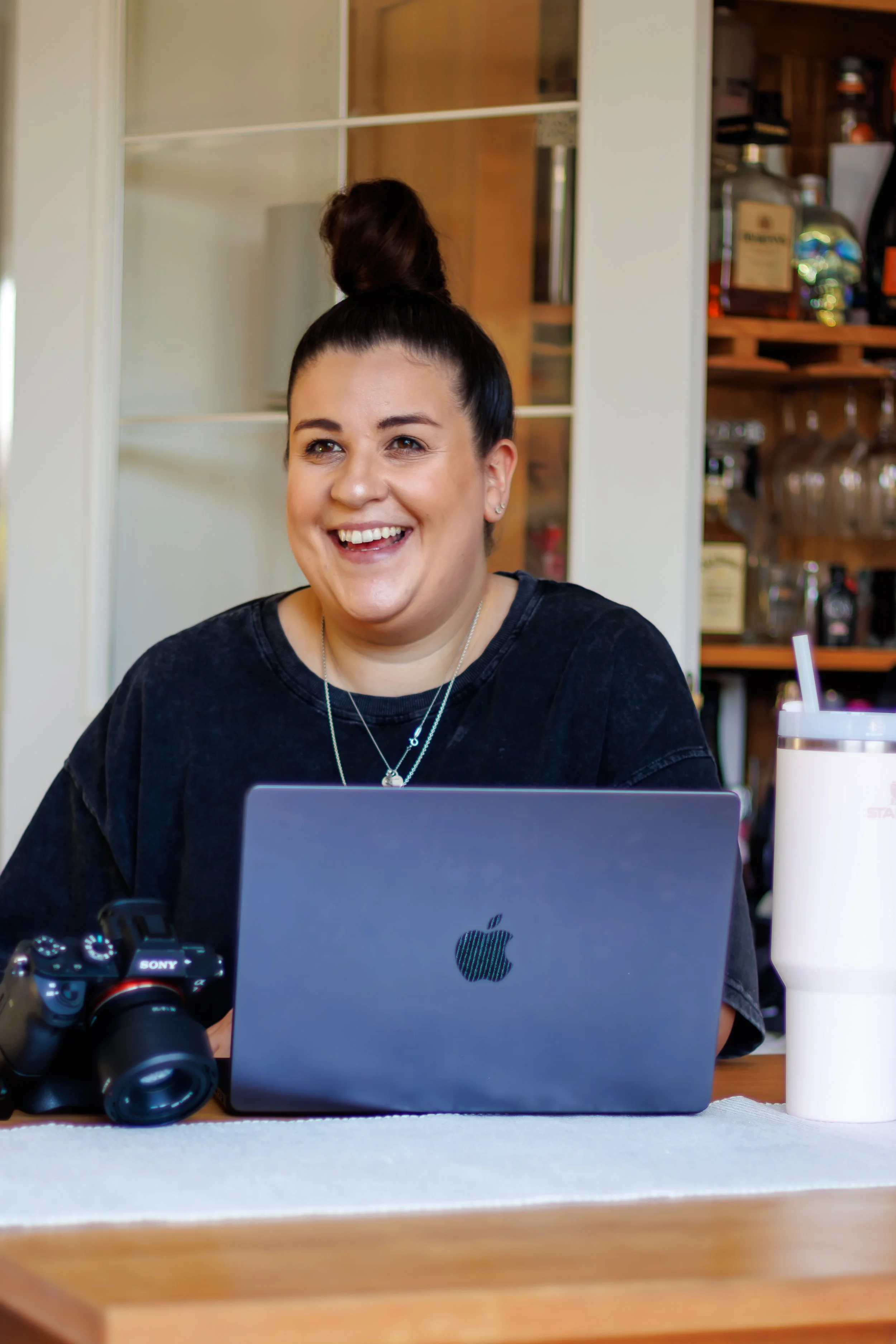 A smiling woman with dark hair in a bun, wearing a black shirt, sitting at a desk with a laptop, camera, and drink, appears to be in a cozy cafe or workspace.