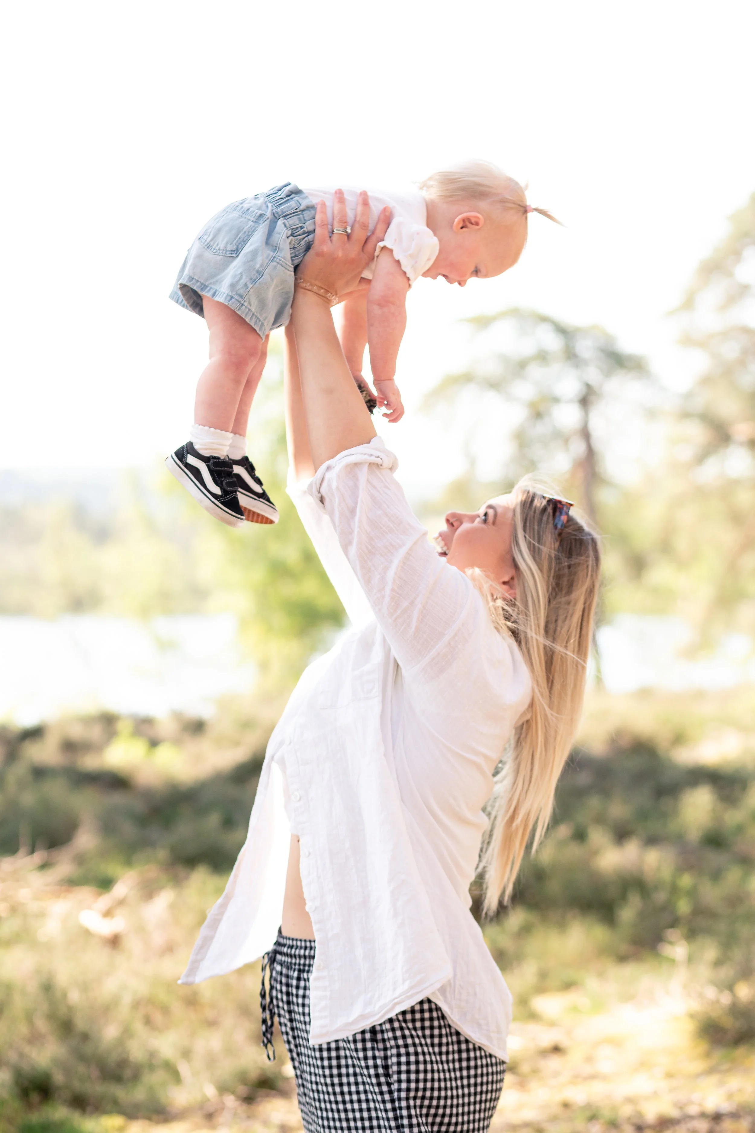 A woman with long blonde hair lifts a smiling young girl high in the air outdoors during daytime, both enjoying a playful moment. - Frensham Little Pond, Surrey.