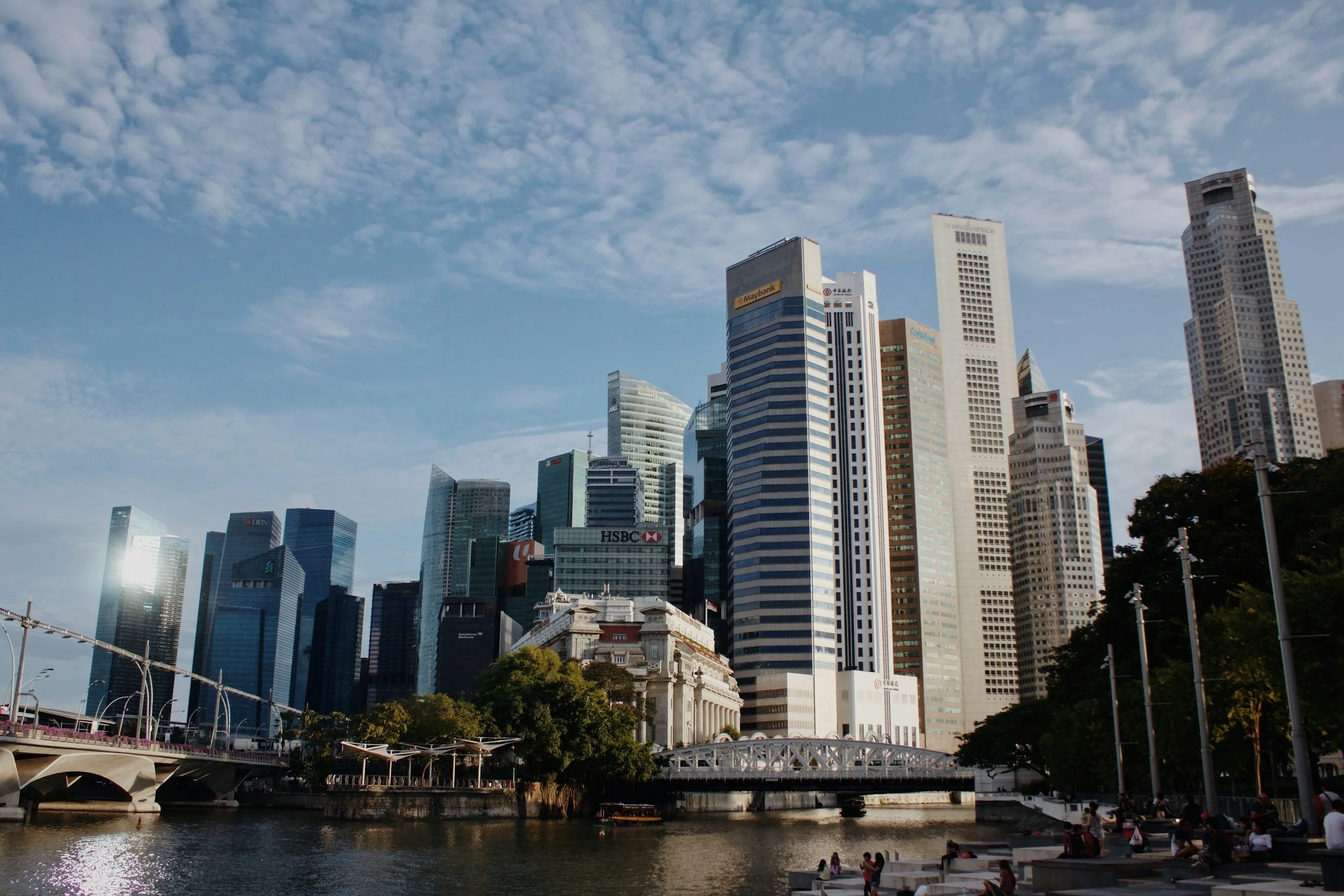 A city skyline with tall modern skyscrapers and a river in the foreground, with people sitting by the riverbank and a bridge crossing the river.