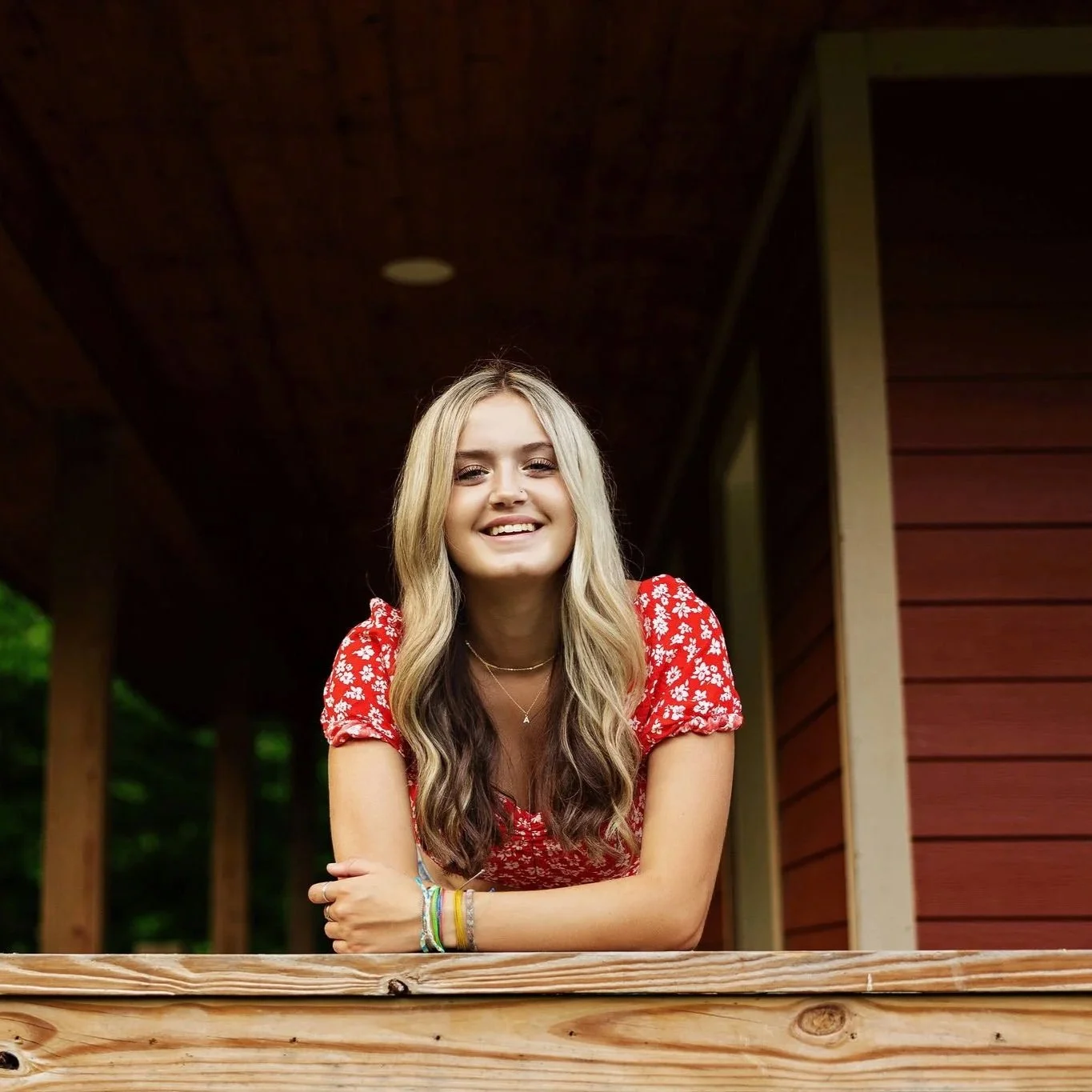 A young woman with long blonde hair smiling, wearing a red floral dress, standing outdoors near a wooden railing with a building and greenery in the background.