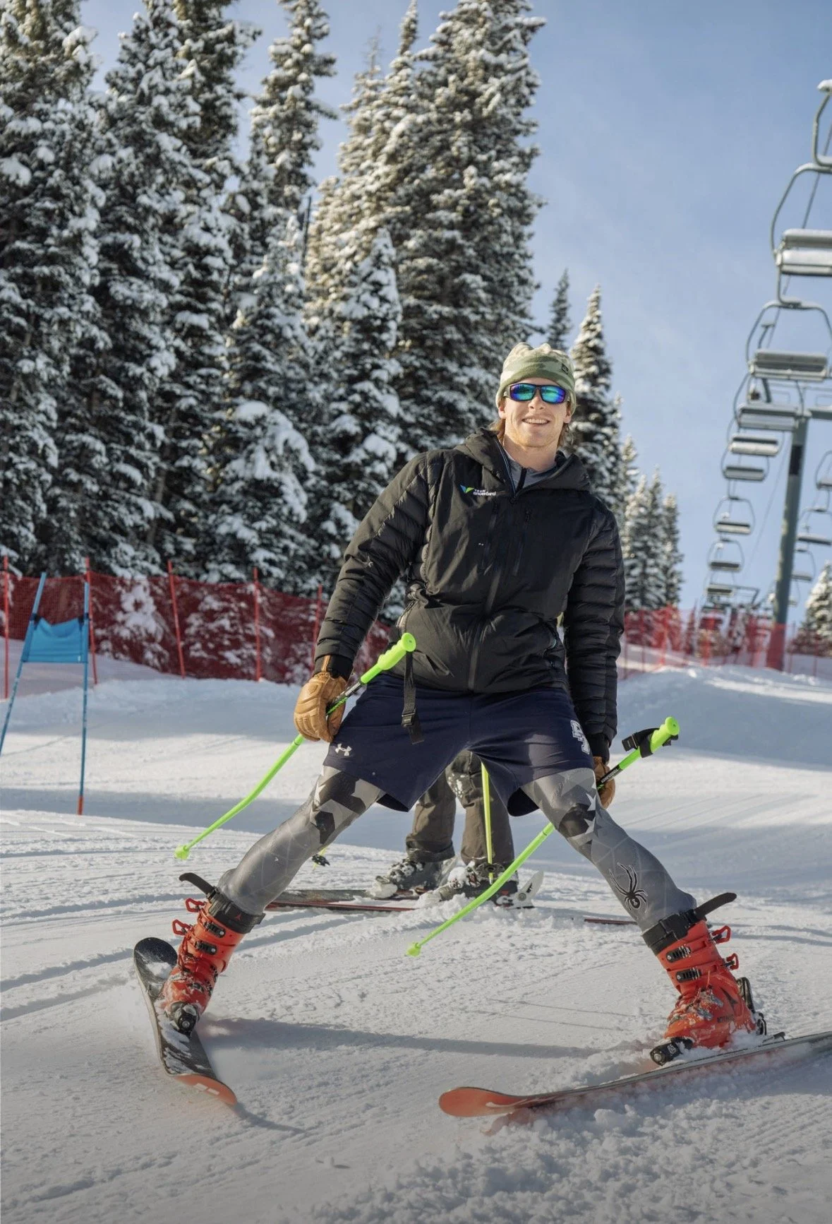 A young man smiling while skiing on snow-covered slopes, wearing sunglasses, a black jacket, and patterned ski pants, with snowy trees and ski lift chairs in the background.