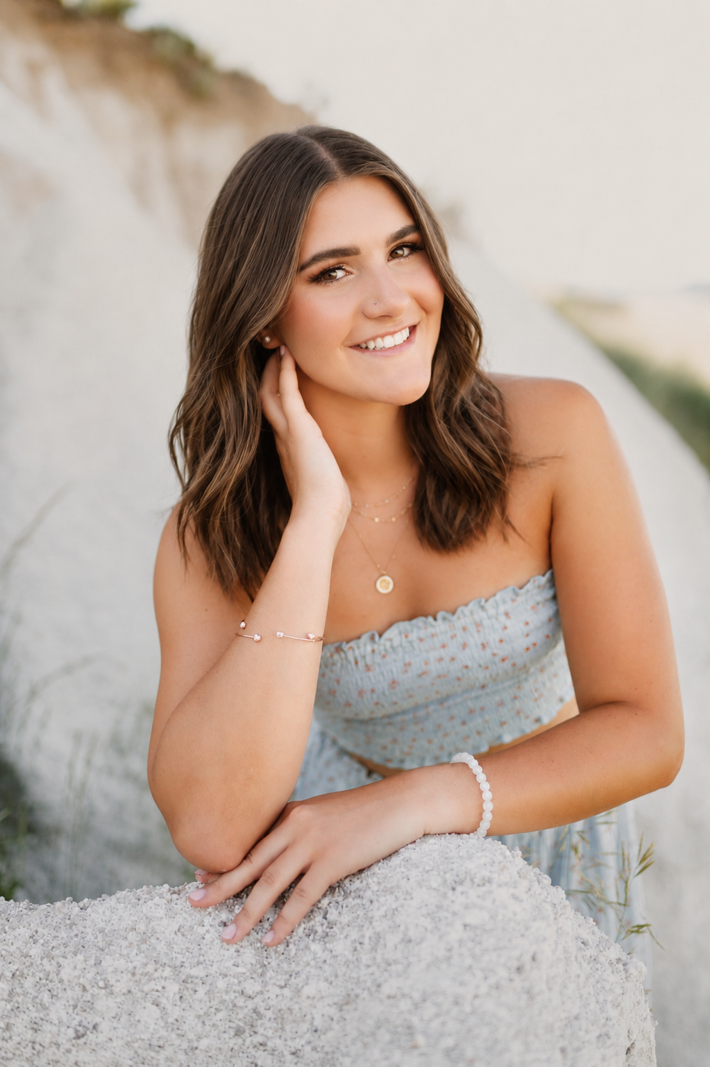 A young woman with long wavy brown hair smiling, sitting outdoors near a sandy area, wearing a light blue strapless dress with small patterns and jewelry.