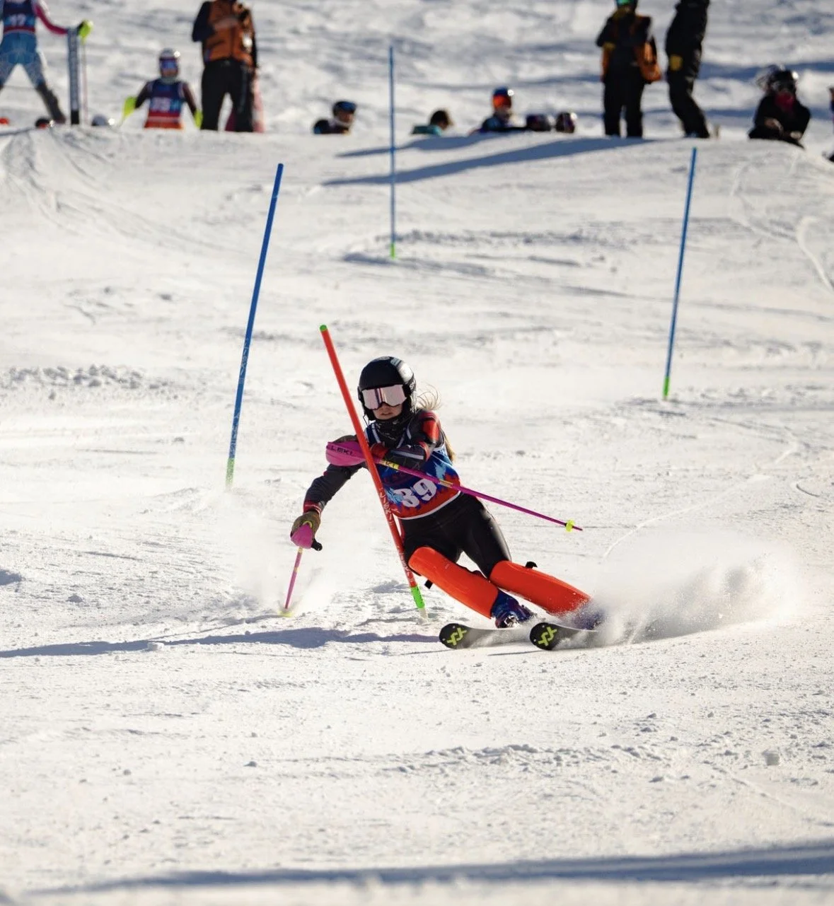 A young skier in a black helmet and goggles navigates through slalom gates on a snowy ski slope during a race or training session.